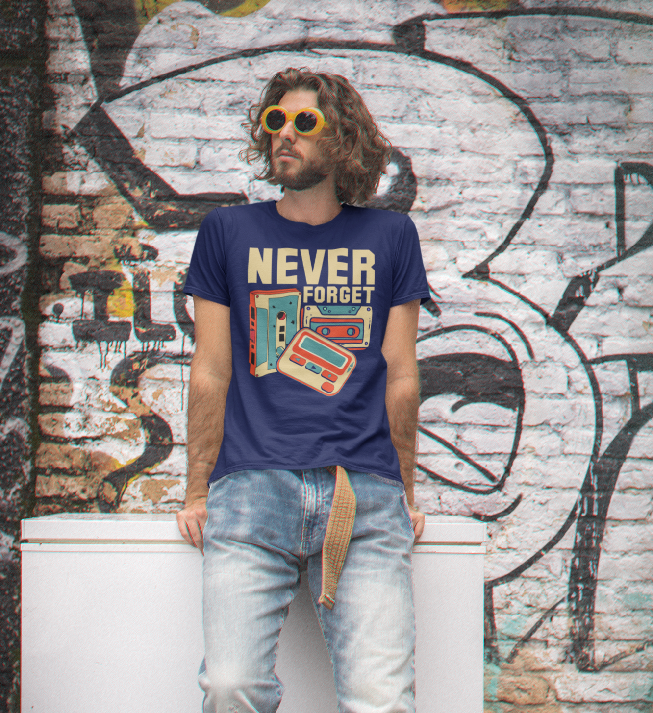 Άντρας με ρετρό εμφάνιση και γυαλιά ηλίου φοράει μπλε μπλούζα με το σύνθημα "Never Forget" και εικόνες από κασέτα, VHS και δισκέτα, μπροστά από γκράφιτι τοίχο.