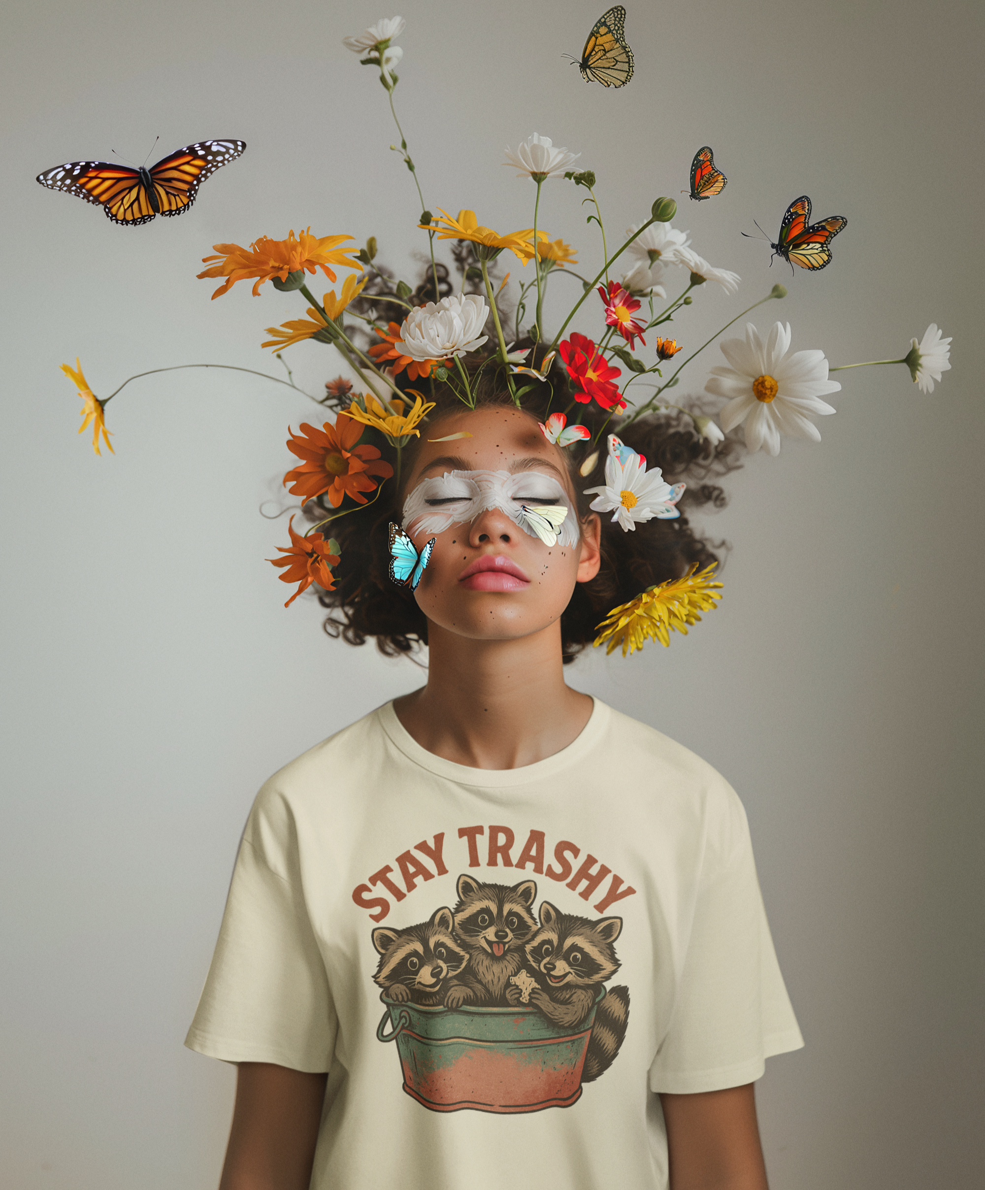 Άτομο με καλλιτεχνικό μακιγιάζ και λουλούδια στο κεφάλι φοράει t-shirt με vintage σχέδιο "Stay Trashy" και τρία ρακούν μέσα σε μεταλλικό κουβά.