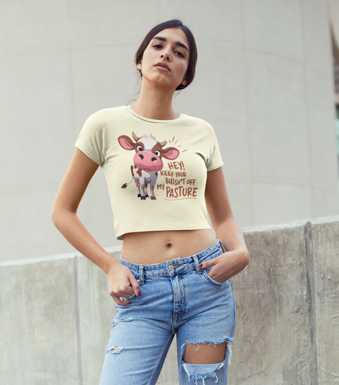 Γυναίκα φοράει ανοιχτόχρωμο crop top με σχέδιο χαριτωμένης αγελάδας και φράση "Hey! Keep your bullsh*t off my pasture", ποζάροντας μπροστά σε μοντέρνο τοίχο.