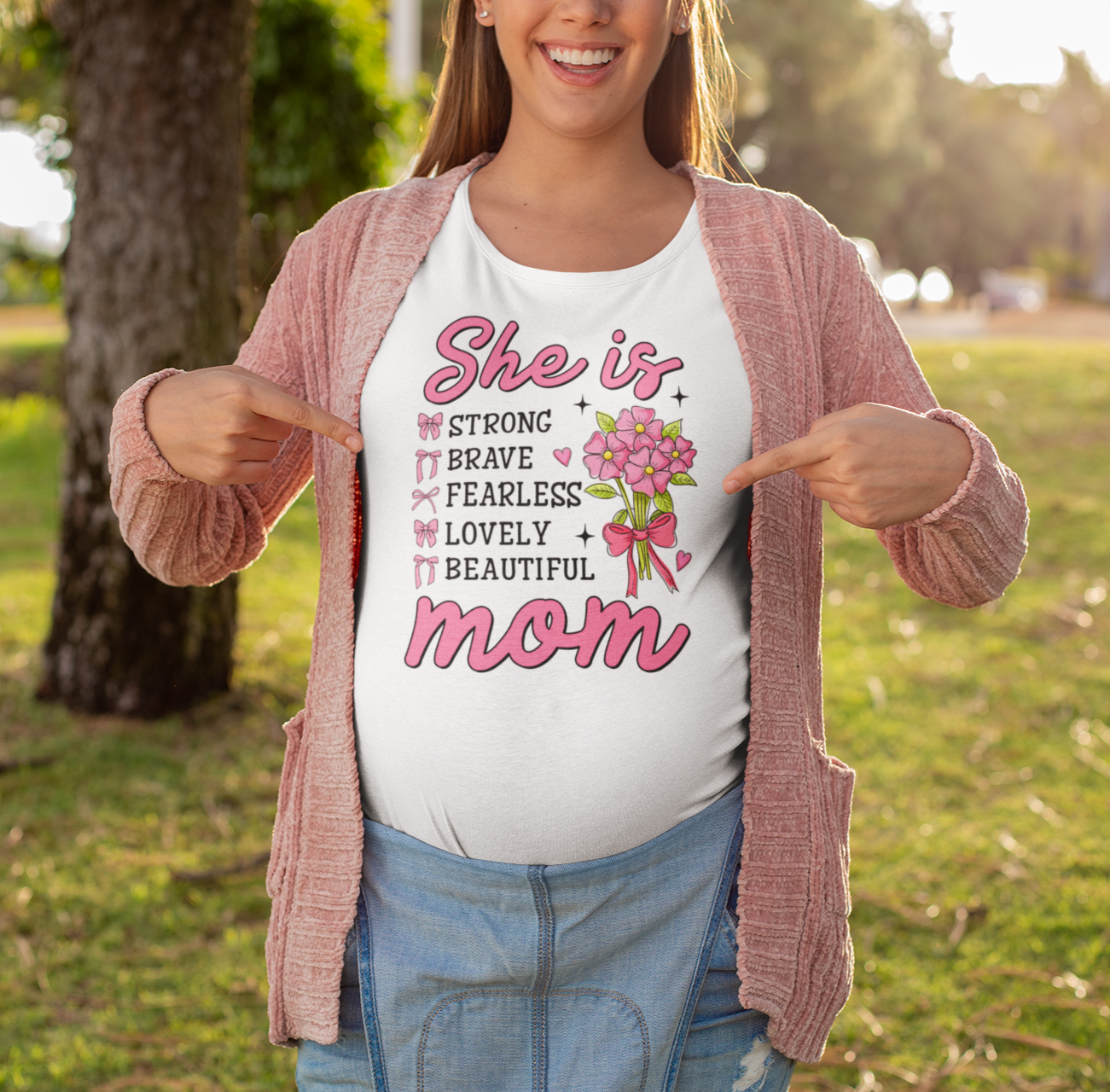λευκό μπλουζάκι που γράφει "She is Mom" με ροζ γράμματα και λουλούδια, ποζάρει χαμογελαστή σε πάρκο δείχνοντας το μπλουζάκι της.