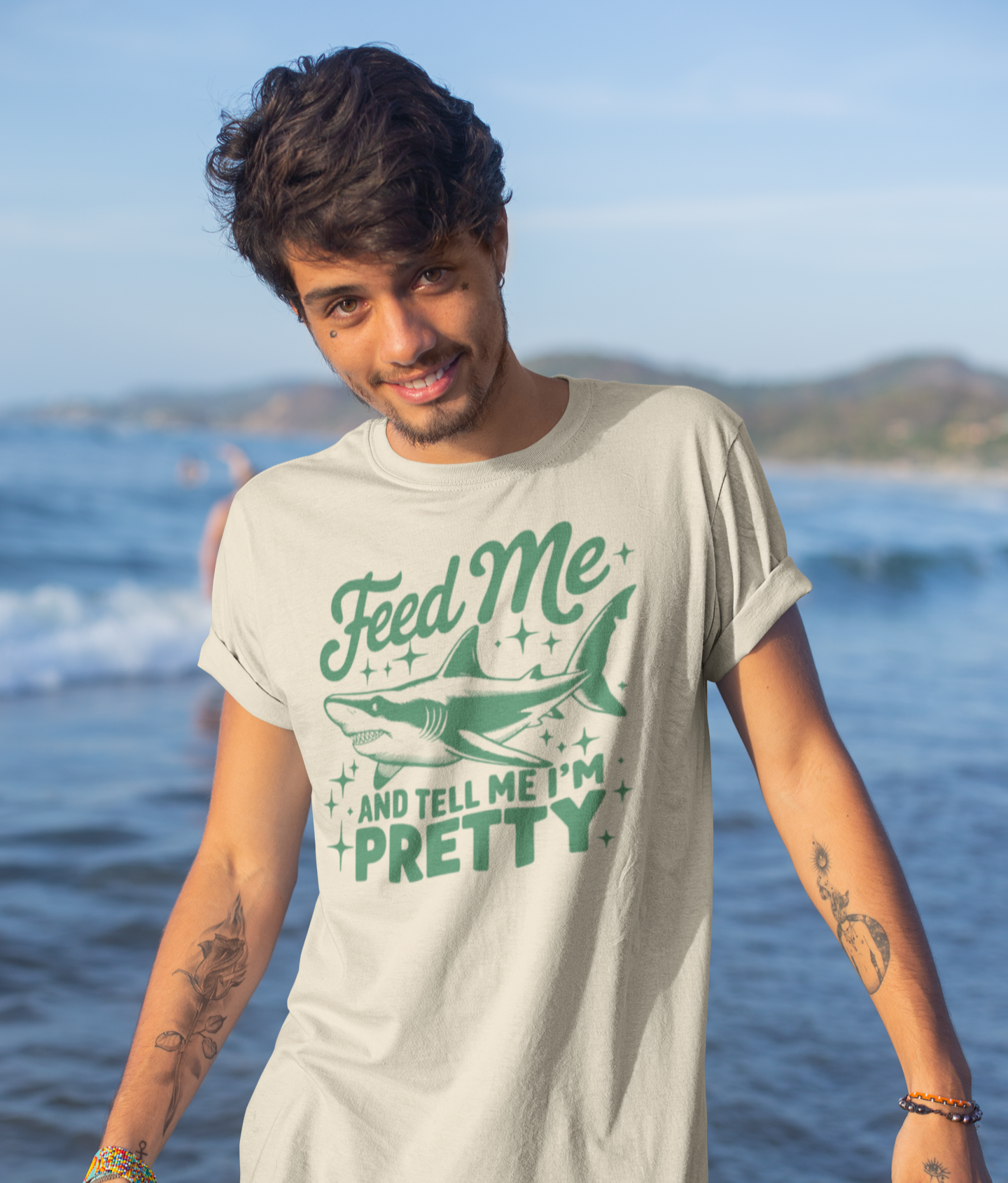 Νεαρός άνδρας με τατουάζ και λευκό μπλουζάκι "Feed Me And Tell Me I’m Pretty" με καρχαρία, ποζάρει μπροστά στη θάλασσα.
