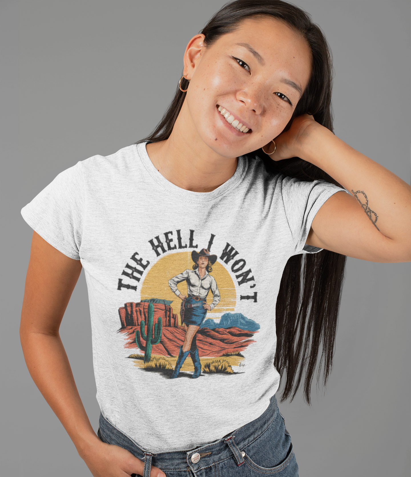 Χαμογελαστή γυναίκα με μακρύ μαλλί φορά γκρι t-shirt με cowgirl γραφικό και το κείμενο “The Hell I Won’t” σε western στυλ.