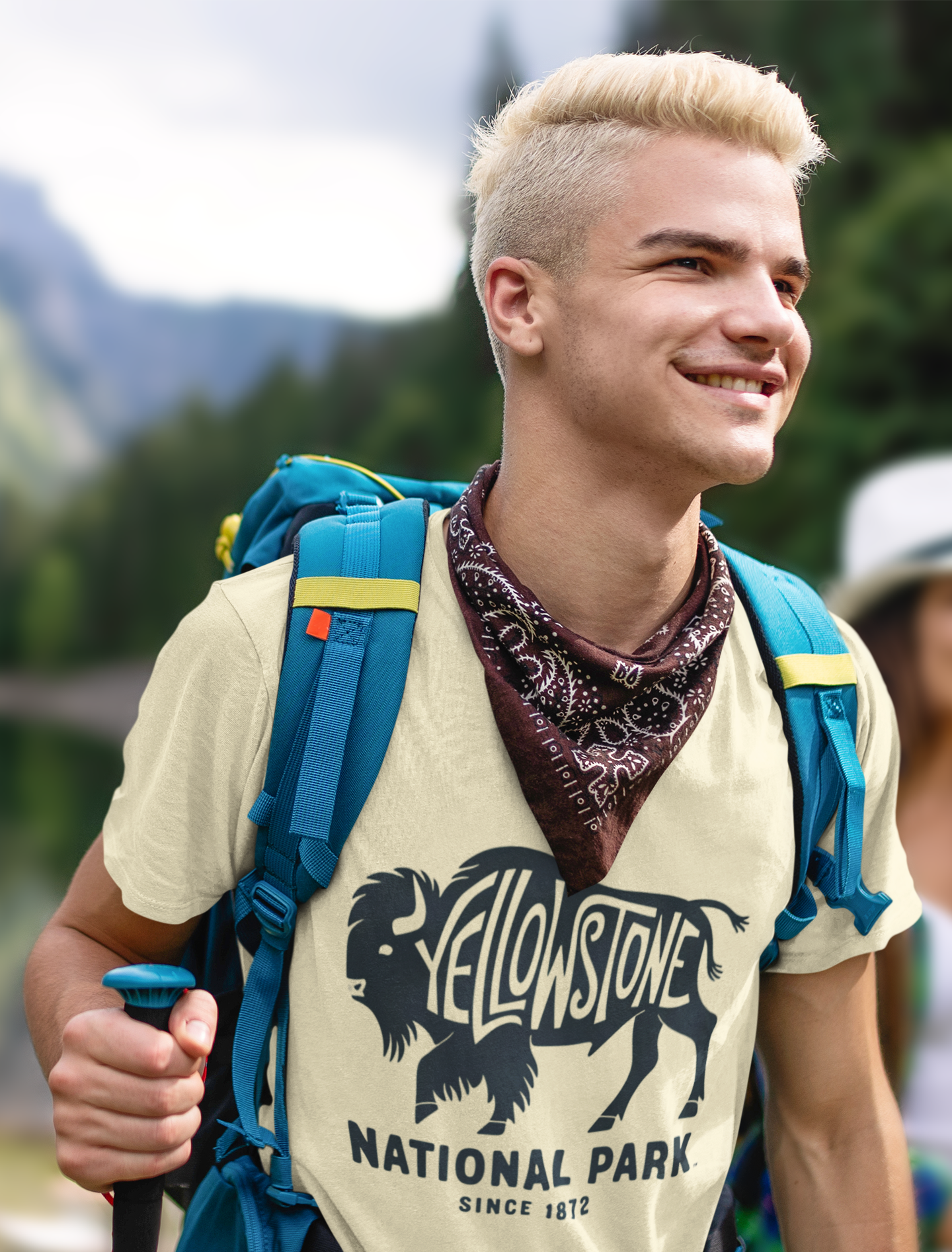 Νεαρός πεζοπόρος με σακίδιο και μπλουζάκι Yellowstone National Park με vintage σχέδιο βίσωνα, σε φυσικό φόντο με δέντρα και λίμνη.