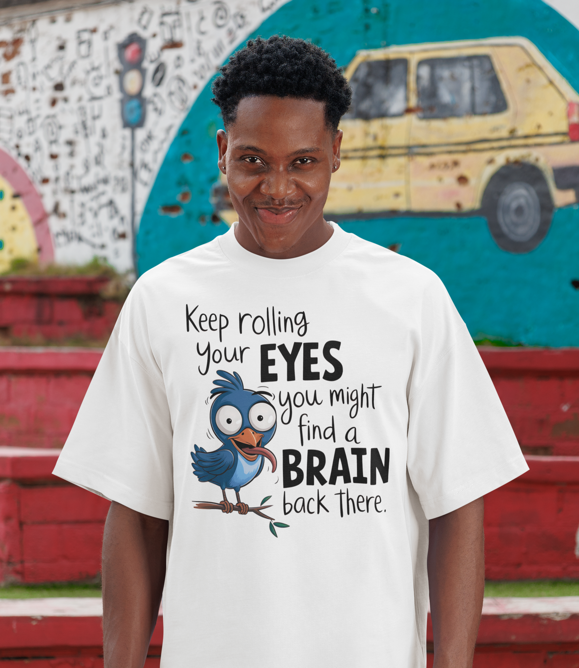 Άντρας με λευκή μπλούζα και αστείο σχέδιο με πουλάκι και τη φράση "Keep rolling your eyes, you might find a brain back there", ποζάρει με χιουμοριστική έκφραση μπροστά σε street art φόντο.