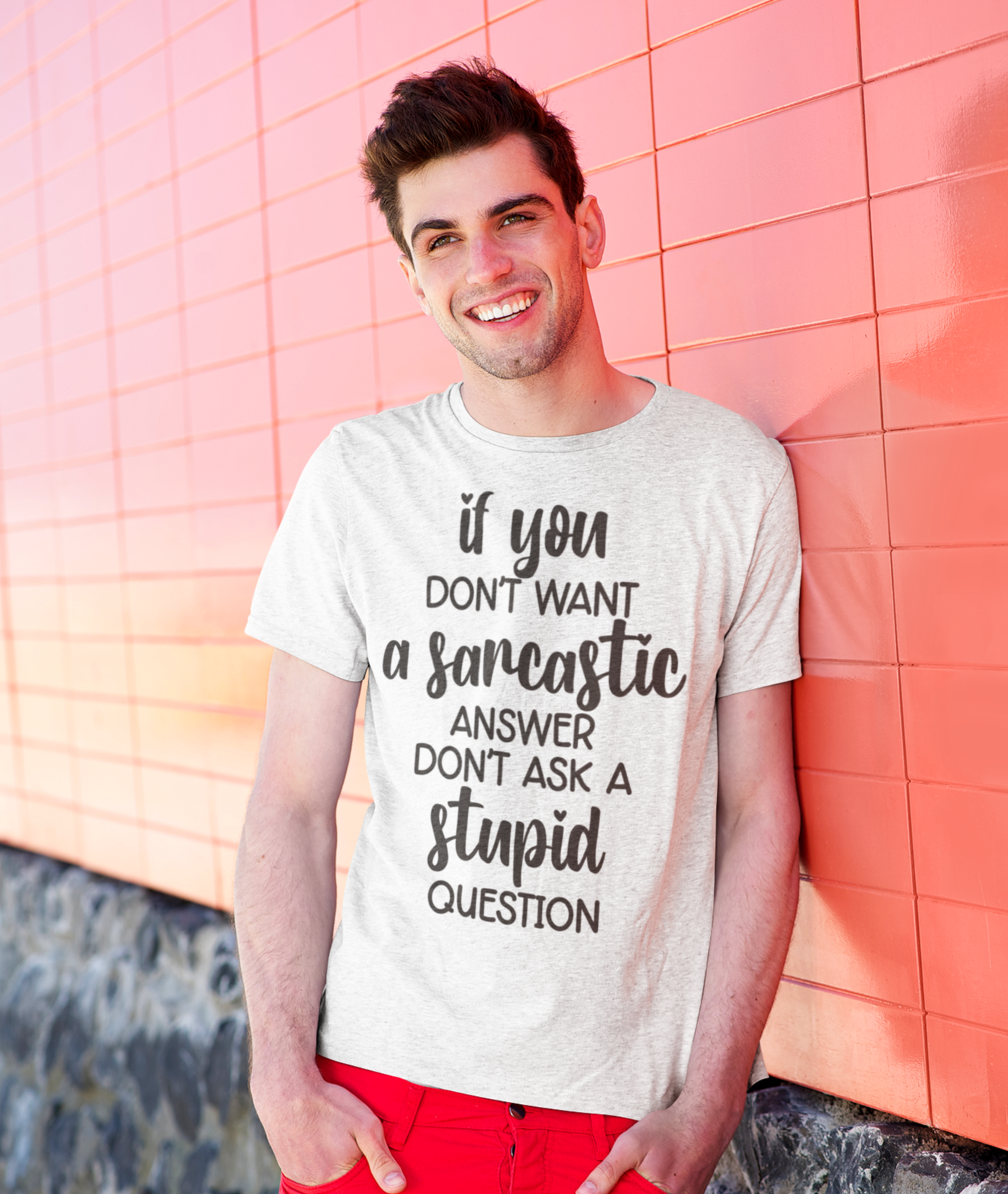 Νεαρός άνδρας χαμογελά φορώντας μπλουζάκι με σαρκαστική φράση, ακουμπώντας σε κόκκινο τοίχο.