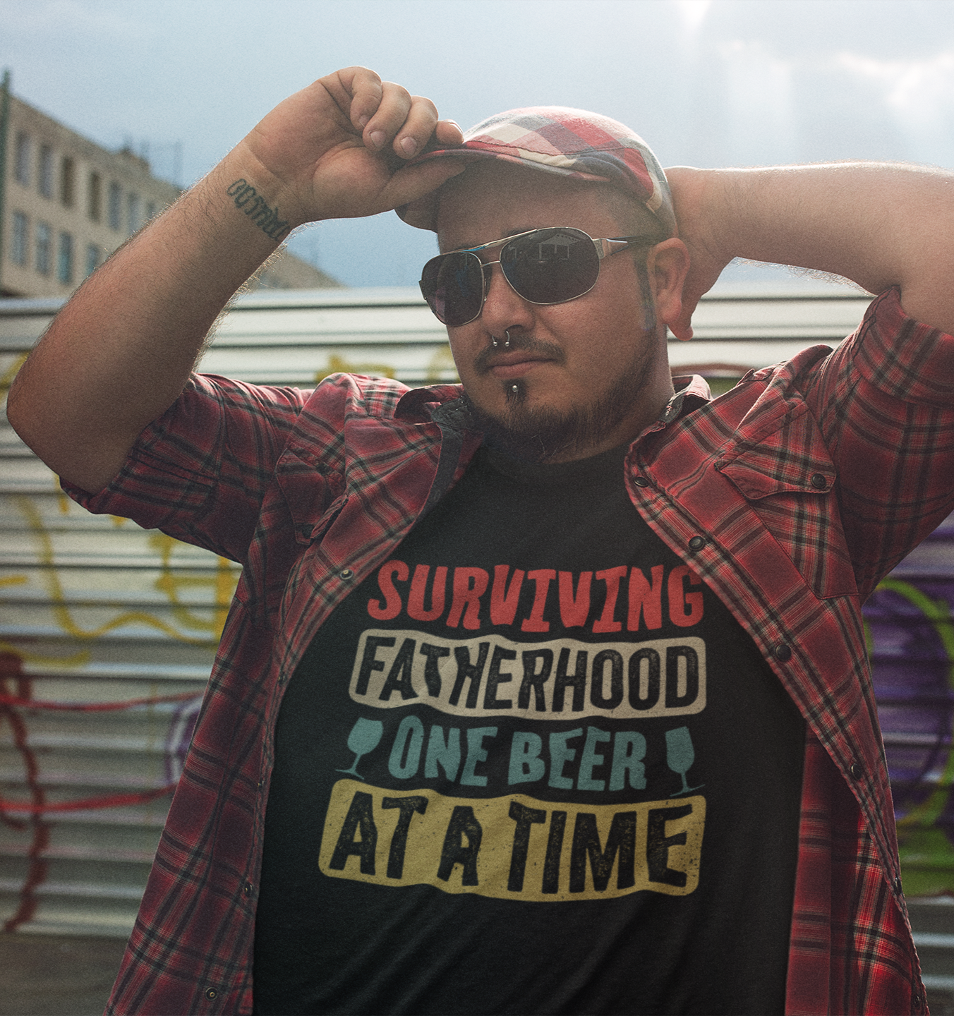 Άντρας με piercing, καρό πουκάμισο και καπέλο φοράει μπλουζάκι “Surviving Fatherhood – One Beer at a Time” σε urban στιλ με πολύχρωμη vintage γραμματοσειρά.