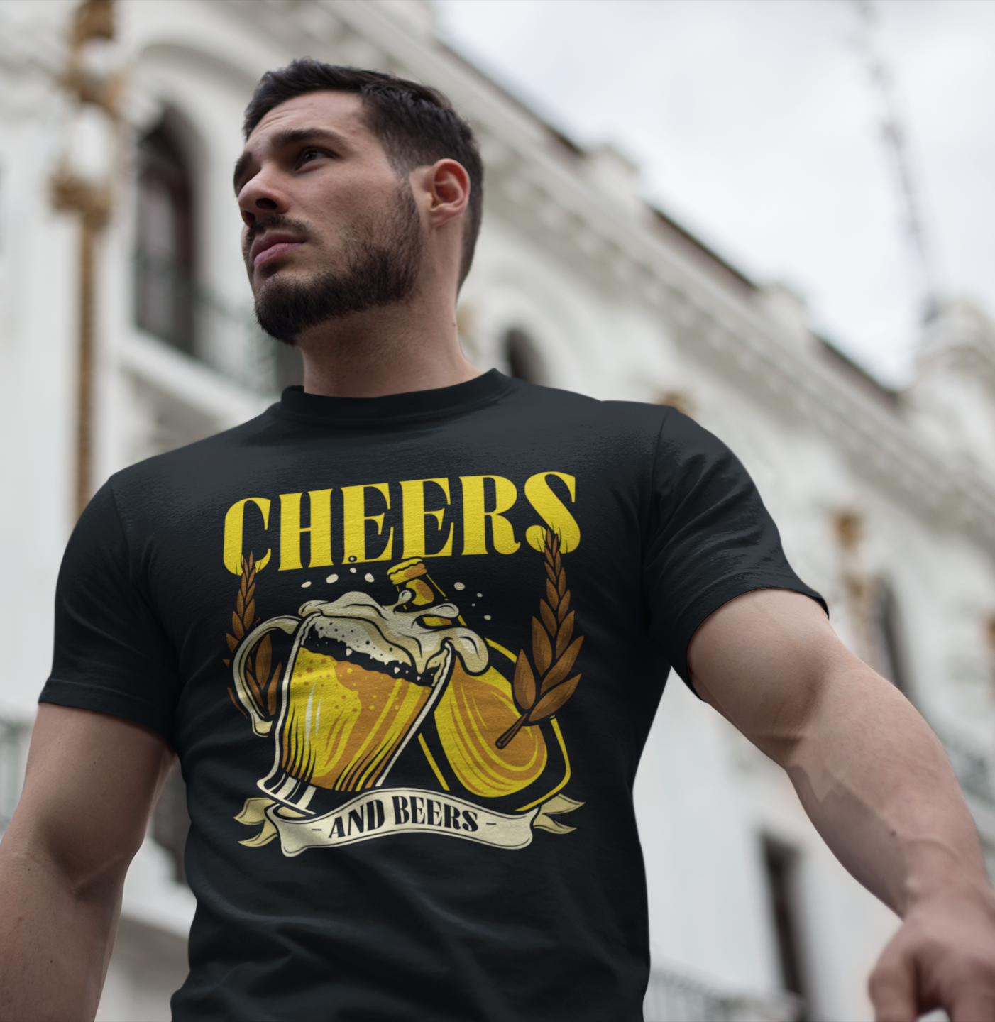 Νεαρός άνδρας φοράει μαύρο t-shirt με στάμπα “Cheers and Beers” και σχέδιο με μπύρες και στάχυα, περπατώντας μπροστά από κλασικό κτίριο στην πόλη.