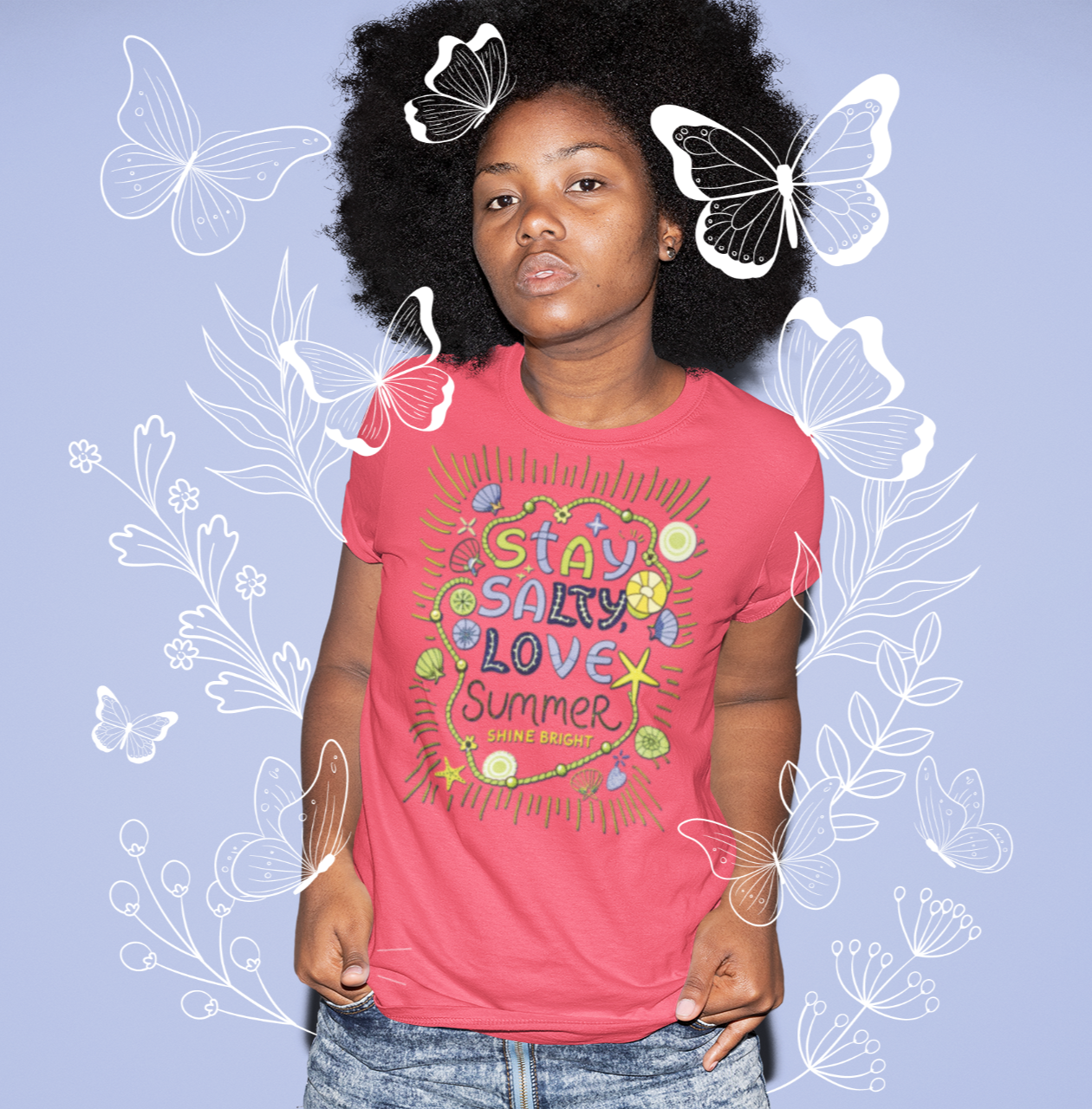 Γυναίκα με φυσικά μαλλιά φοράει κόκκινο μπλουζάκι με πολύχρωμο σχέδιο "Stay Salty Love Summer", μπροστά από μωβ φόντο με λευκές πεταλούδες και λουλούδια.