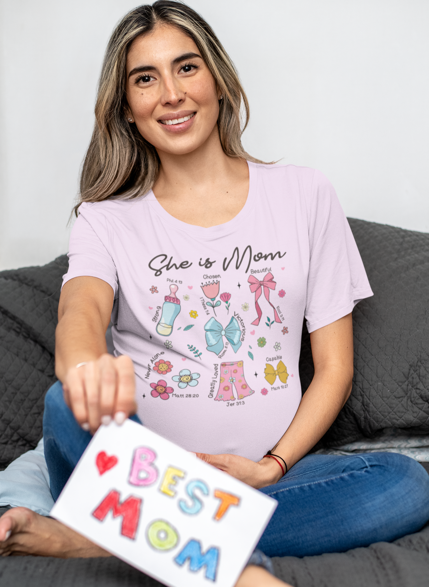 Γυναίκα με ανοιχτό ροζ μπλουζάκι που γράφει "She is Mom" και κρατά ζωγραφισμένη κάρτα "Best Mom", καθισμένη σε γκρι καναπέ.