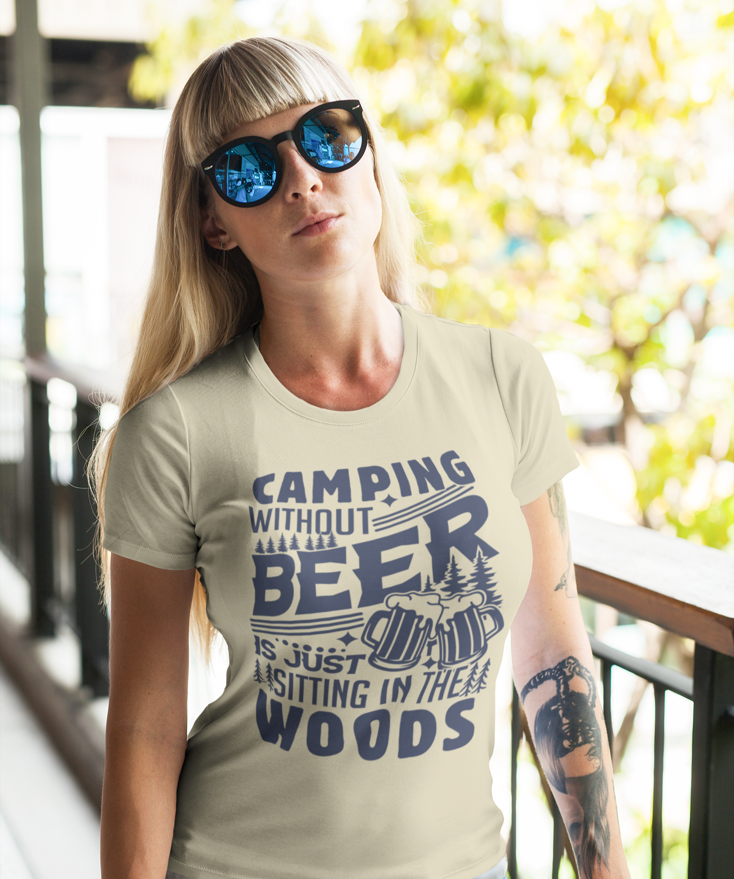 Γυναίκα με μακριά ξανθά μαλλιά και γυαλιά ηλίου φοράει μπεζ t-shirt με στάμπα “Camping Without Beer Is Just Sitting In The Woods”, ποζάροντας σε εξωτερικό χώρο με φόντο δέντρα.