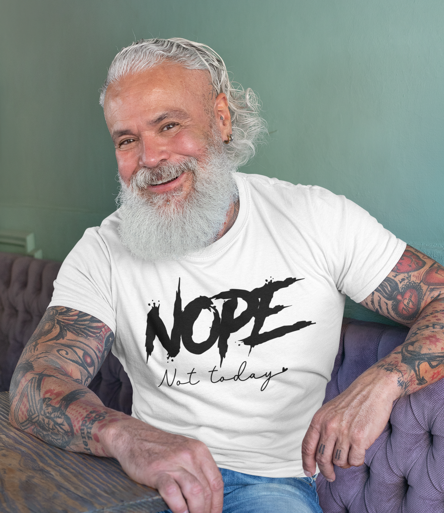 Γοητευτικός τατουαρισμένος ηλικιωμένος άνδρας με γένια φοράει λευκό μπλουζάκι με στάμπα “NOPE Not Today” και χαμογελάει ενώ κάθεται σε καφετέρια.