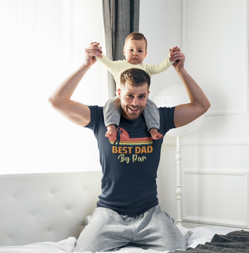Μπαμπάς με μπλουζάκι "Best Dad by Par" κάθεται στο κρεβάτι και κρατάει το παιδί του στους ώμους του, σε φωτεινό υπνοδωμάτιο
