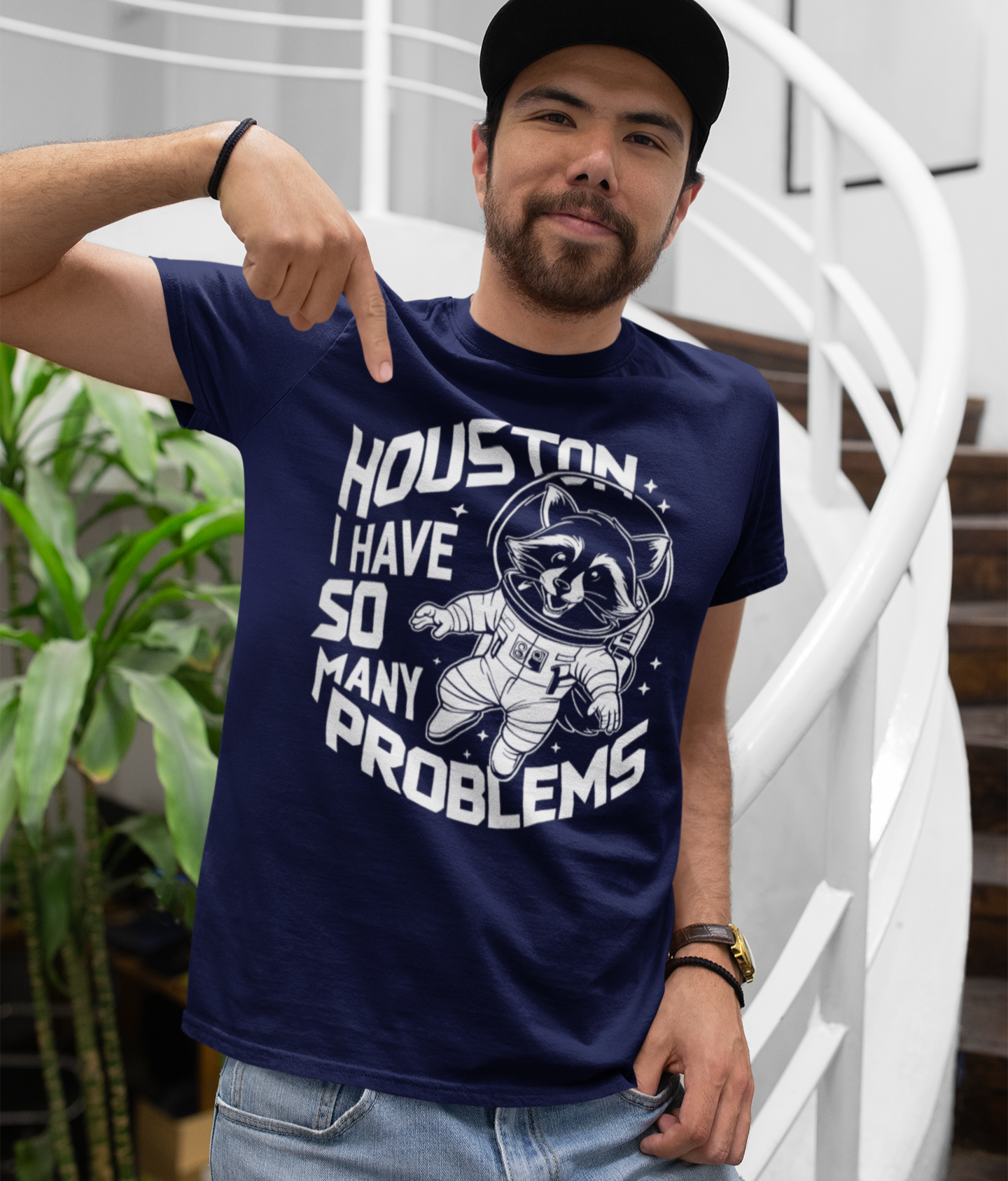 Νεαρός άνδρας με καπέλο δείχνει το μπλουζάκι του που γράφει “Houston I have so many problems” με σχέδιο ρακούν-αστροναύτη.