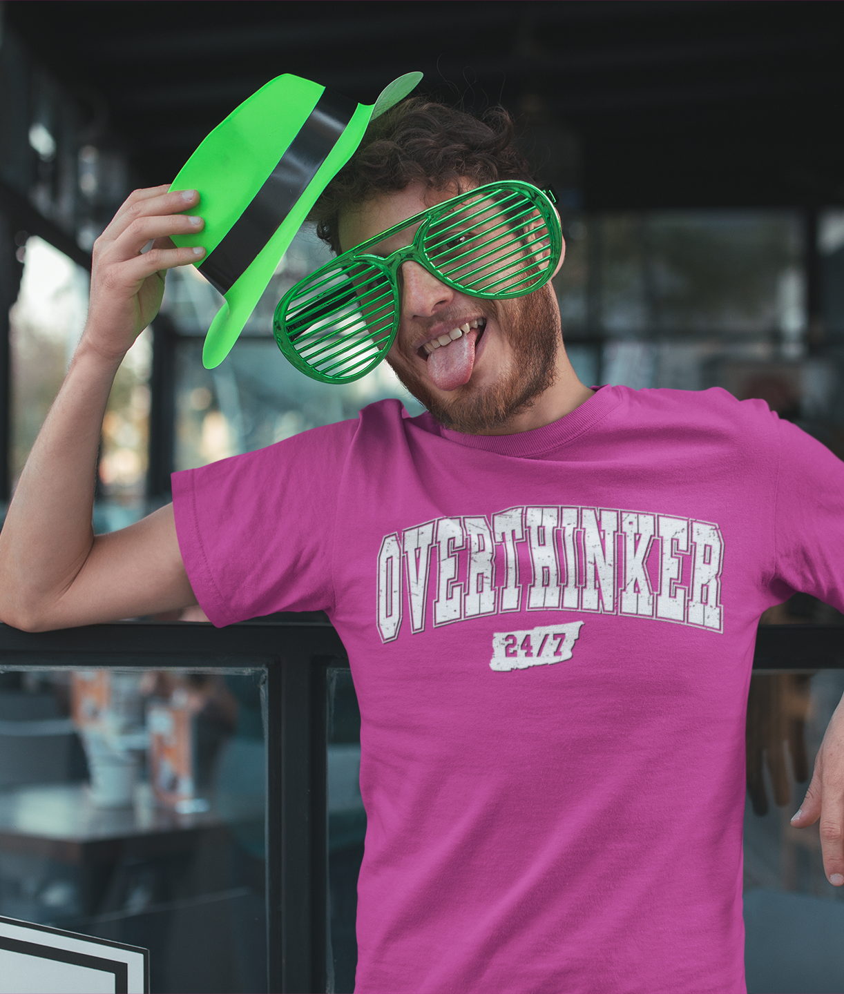 Νεαρός άνδρας με πράσινα αστεία γυαλιά και καπέλο του Αγίου Πατρικίου φοράει ροζ μπλουζάκι με στάμπα “OVERTHINKER 24/7” και βγάζει τη γλώσσα με παιχνιδιάρικη διάθεση.