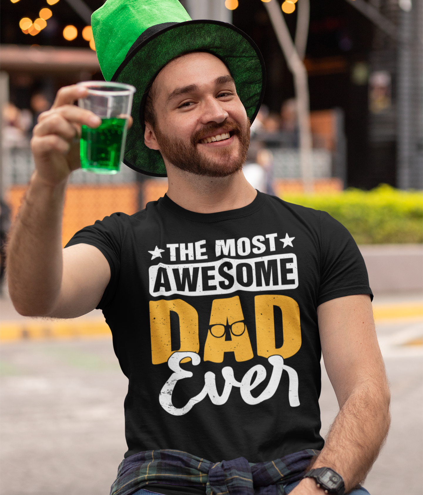 Χαμογελαστός άντρας με πράσινο καπέλο κρατάει ποτήρι με πράσινη μπύρα και φοράει μαύρο μπλουζάκι με το μήνυμα “The Most Awesome Dad Ever”.