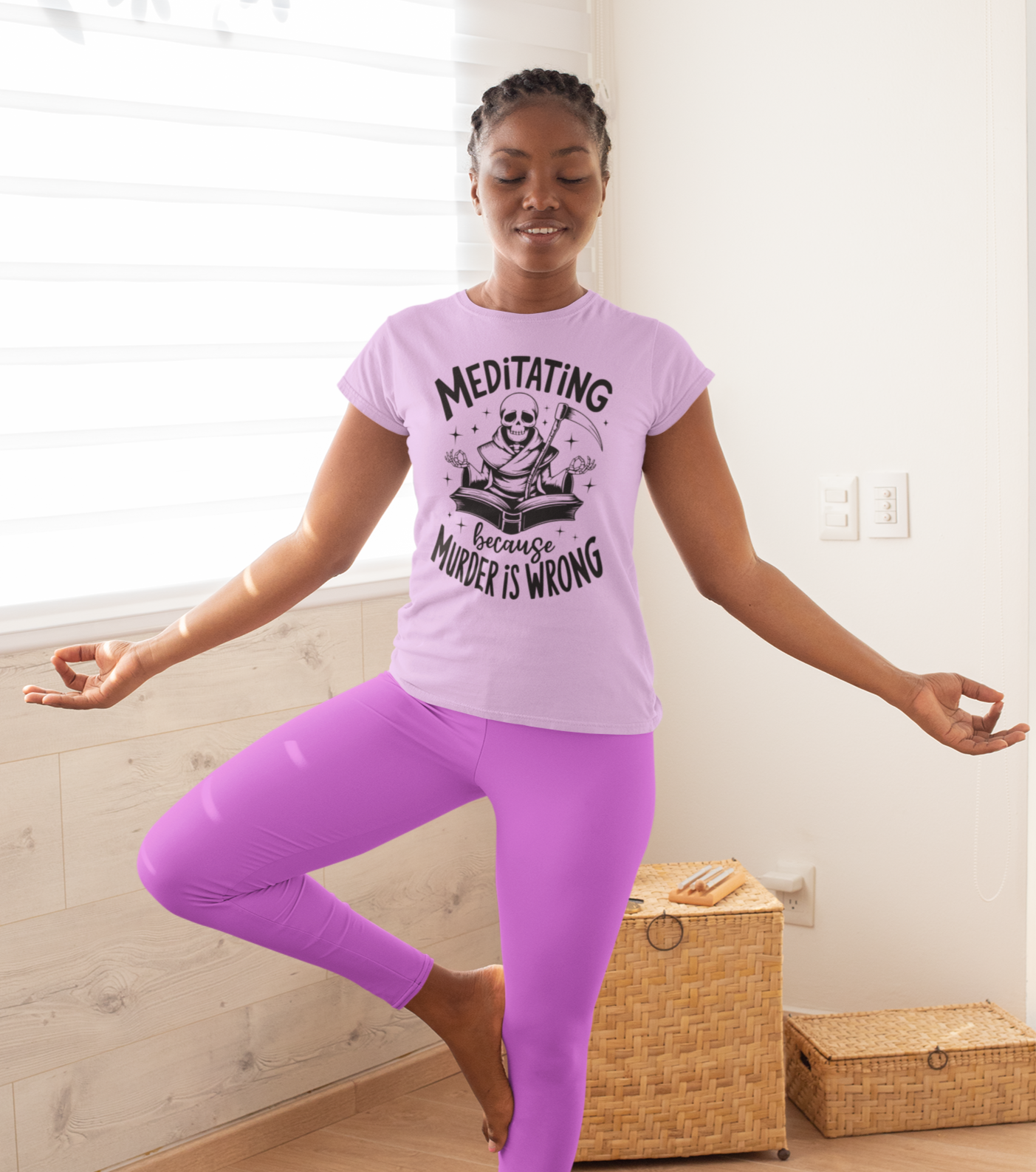 Γυναίκα με λιλά μπλουζάκι και κολάν κάνει yoga πόζα σε φωτεινό δωμάτιο. Το μπλουζάκι γράφει “Meditating because murder is wrong” με σχέδιο σκελετού σε στάση λωτού.