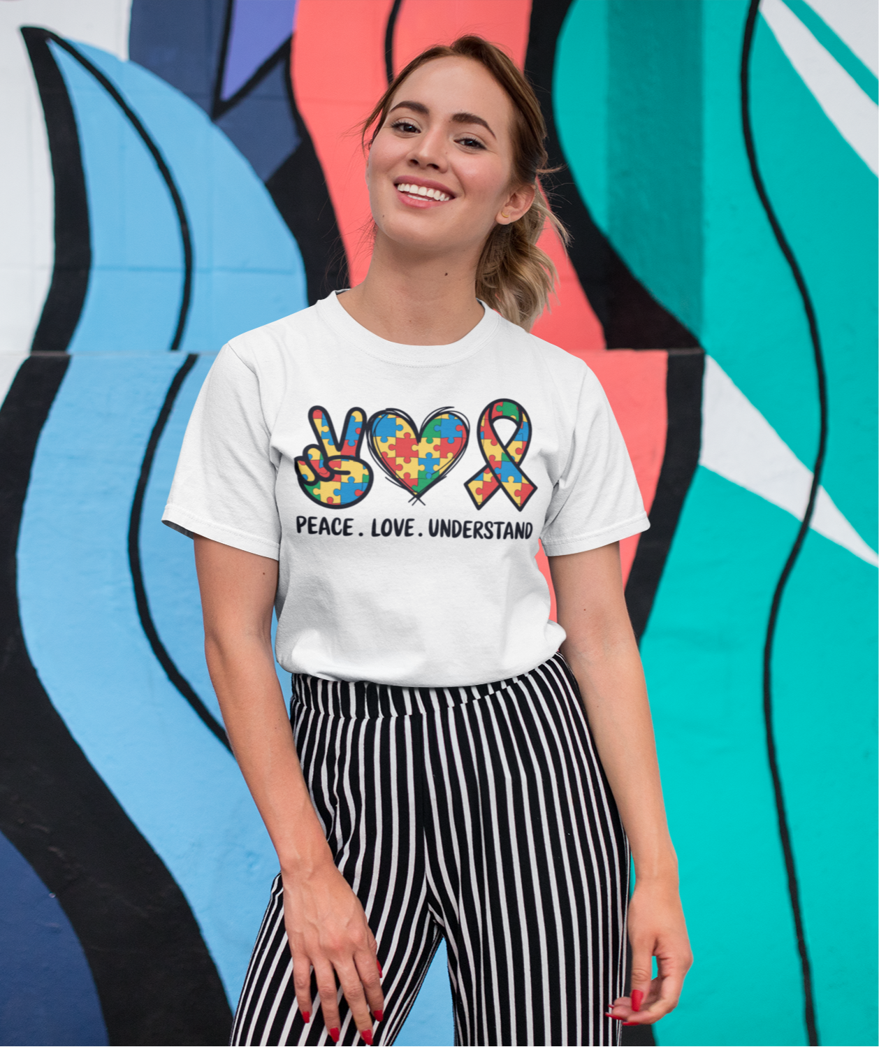λευκό μπλουζάκι που γράφει "Peace. Love. Understand." με πολύχρωμα σύμβολα αυτισμού, ποζάρει χαμογελαστή μπροστά σε τοίχο με πολύχρωμο γκράφιτι.