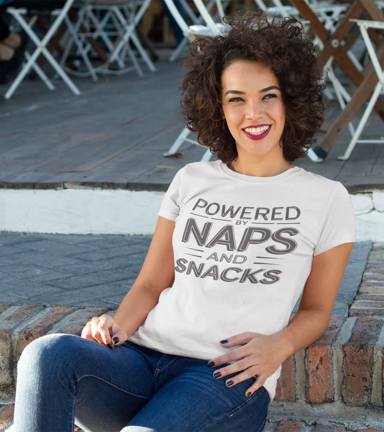 Γυναίκα με σγουρά μαλλιά κάθεται χαμογελαστή σε εξωτερικό χώρο φορώντας λευκό μπλουζάκι με στάμπα “Powered by Naps and Snacks”.