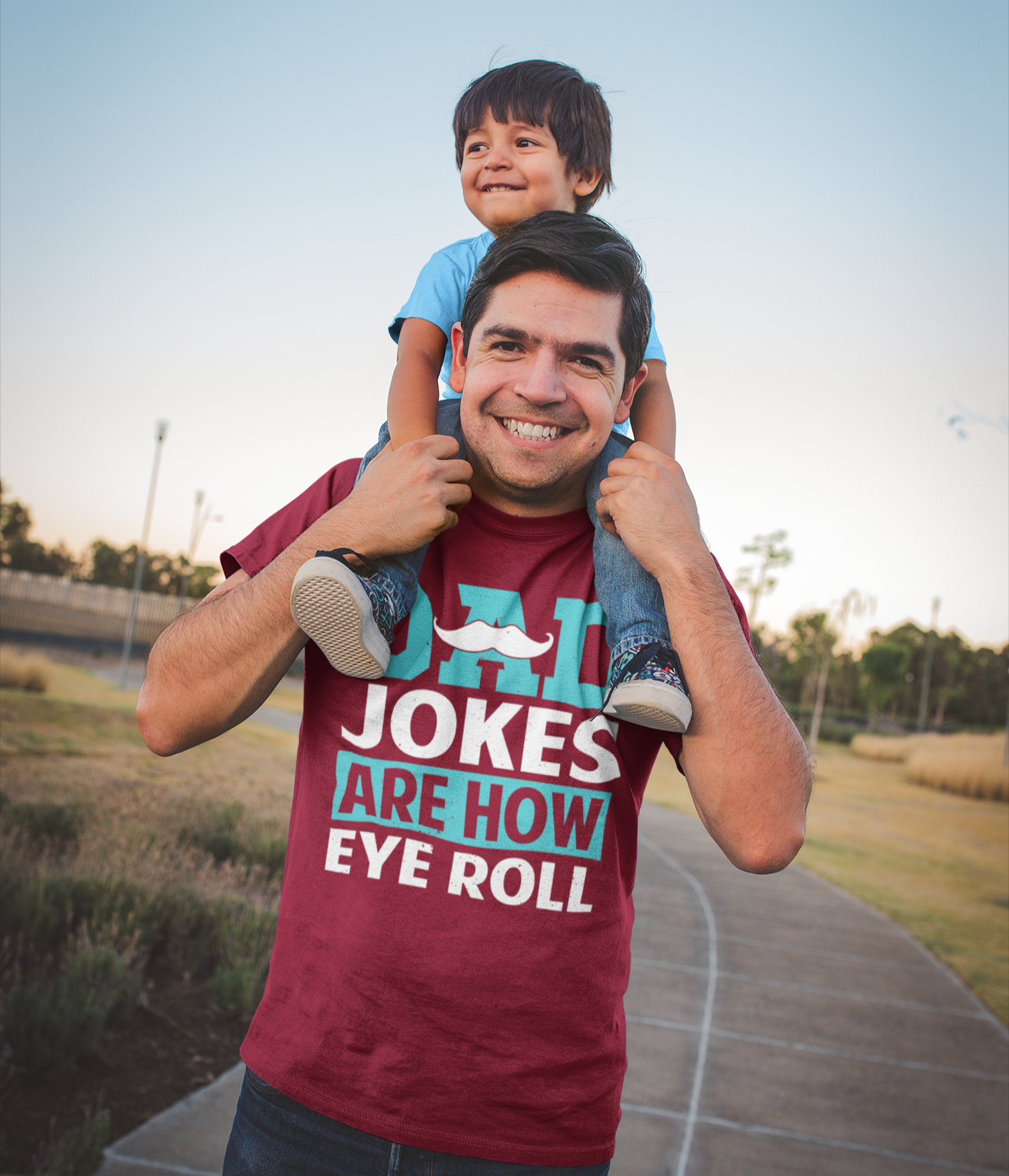 Μπαμπάς που φοράει μπλουζάκι με στάμπα “Dad Jokes Are How I Eye Roll” και κουβαλάει τον γιο του στους ώμους σε πάρκο