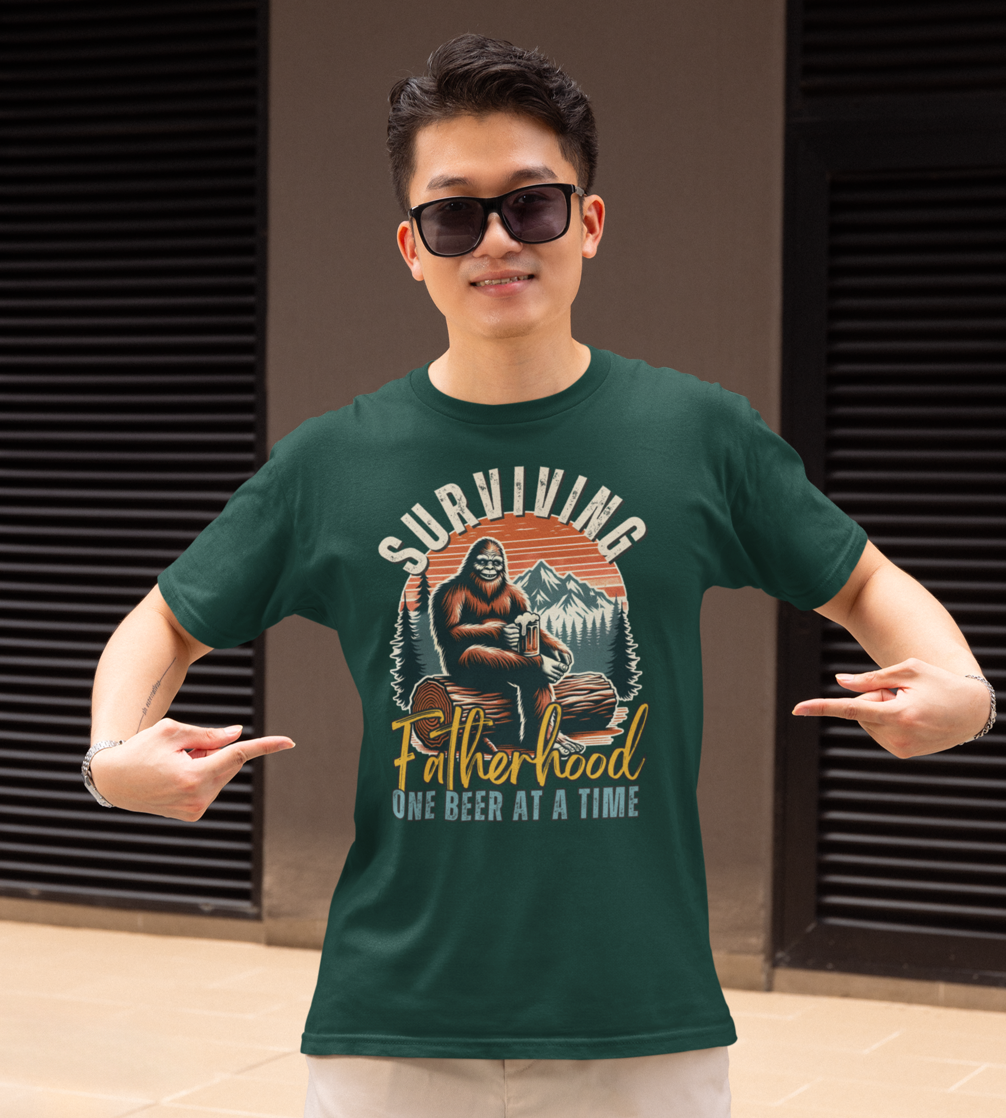 Νεαρός άνδρας δείχνει χαμογελαστός το πράσινο μπλουζάκι του με στάμπα Bigfoot και τη φράση “Surviving Fatherhood One Beer at a Time”, σε εξωτερικό χώρο.