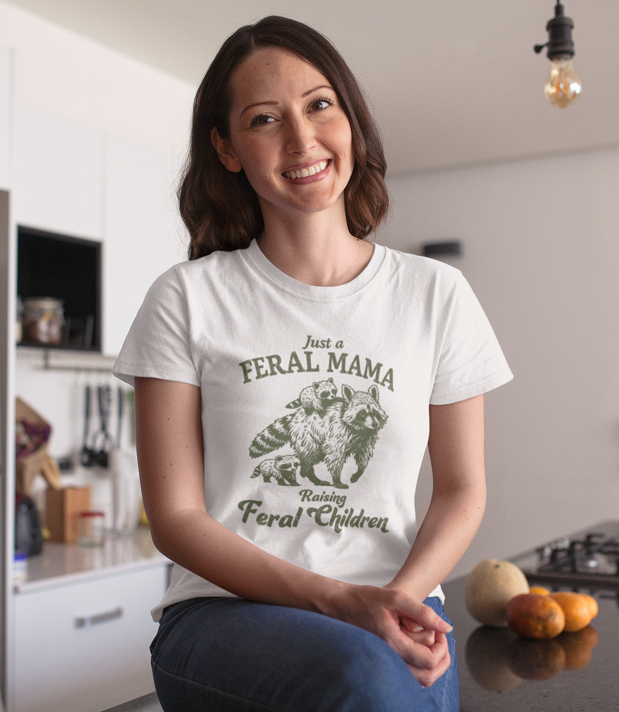 Γυναίκα με λευκό μπλουζάκι που γράφει "Just a Feral Mama Raising Feral Children" με σχέδιο ρακούν, χαμογελάει καθισμένη σε κουζίνα.