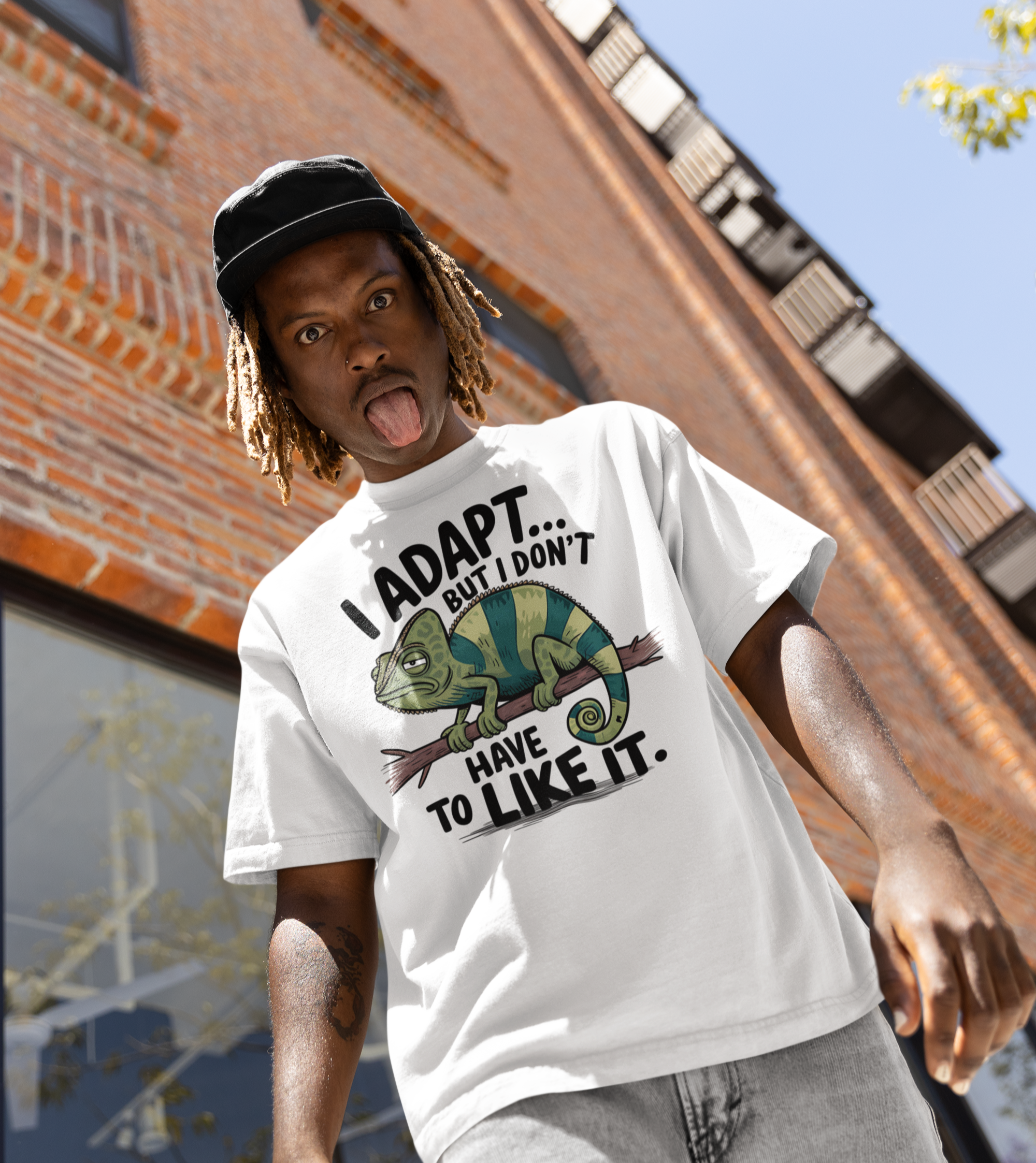 Νεαρός άνδρας με λευκό t-shirt με σχέδιο χαμαιλέοντα και φράση “I Adapt But I Don’t Have to Like It” μπροστά από κτήριο τούβλου