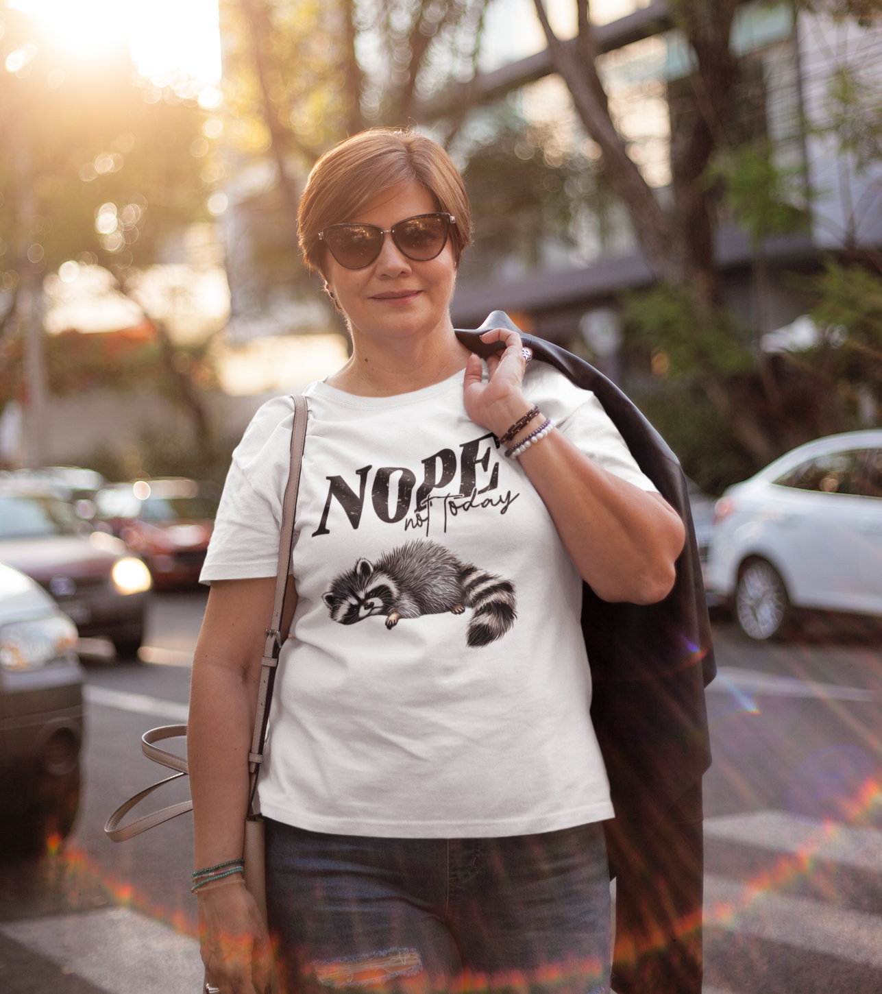 Γυναίκα μέσης ηλικίας φοράει λευκό μπλουζάκι με στάμπα ρακούν και τη φράση “Nope Not Today” ενώ περπατάει χαλαρά στον δρόμο, στο ηλιοβασίλεμα.