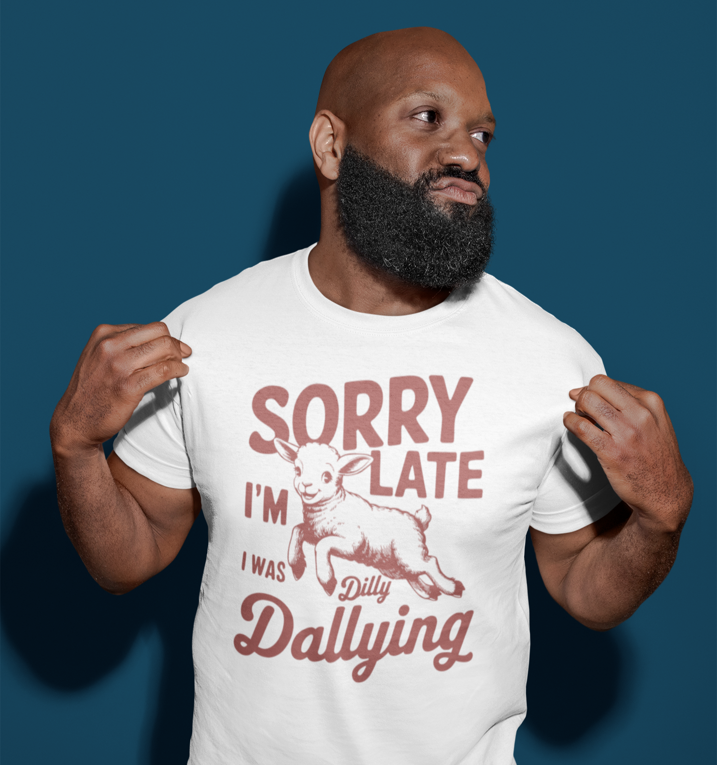 Άντρας με λευκό μπλουζάκι που γράφει "Sorry I’m Late I Was Dilly Dallying" και απεικονίζει ένα χαριτωμένο αρνάκι, ποζάρει κρατώντας τη μπλούζα του με φόντο μπλε τοίχο.