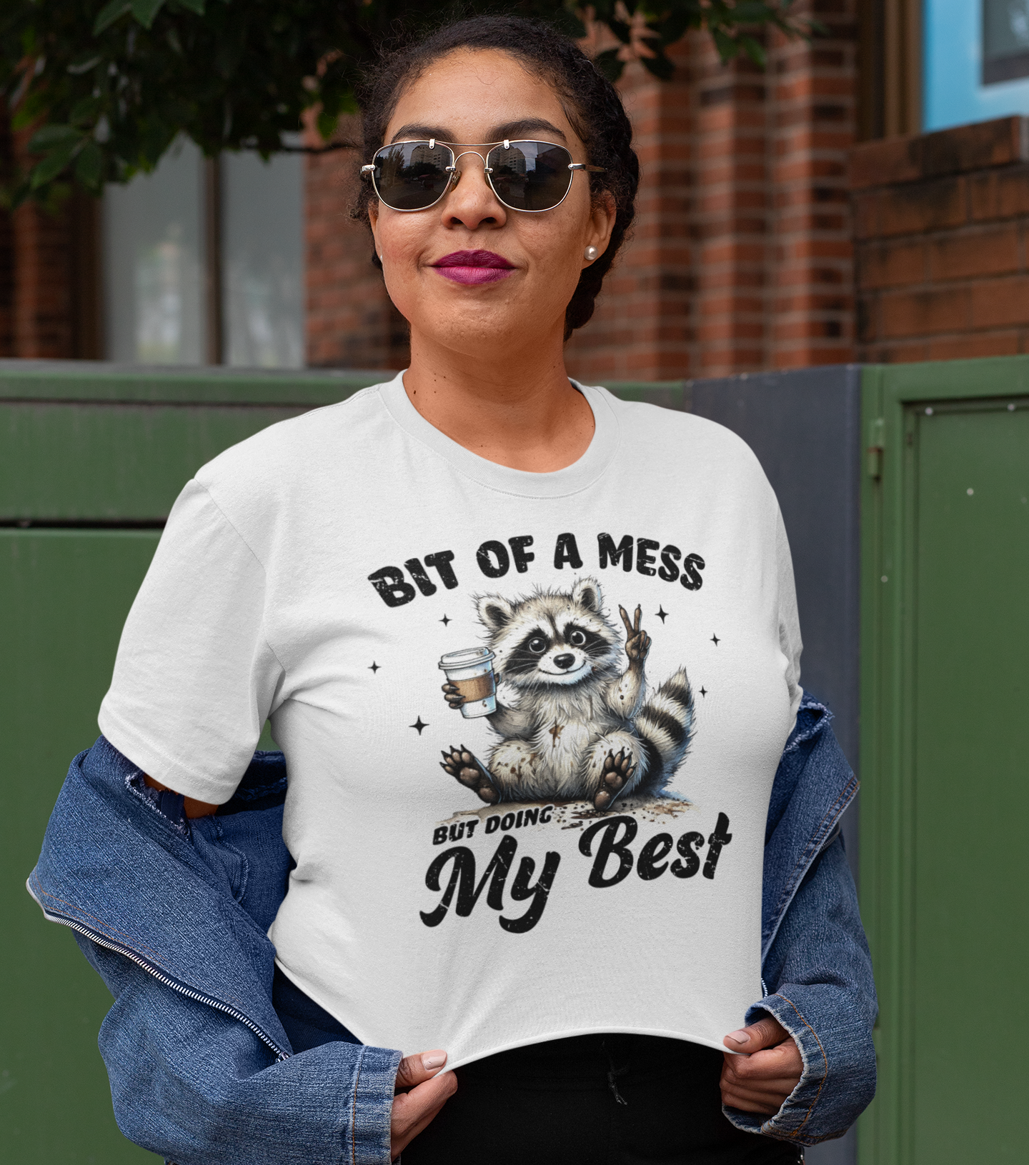 Γυναίκα φορά λευκό crop top με σχέδιο ρακούν που κρατά καφέ και γράφει "Bit of a Mess but Doing My Best", σε αστικό περιβάλλον με casual outfit.