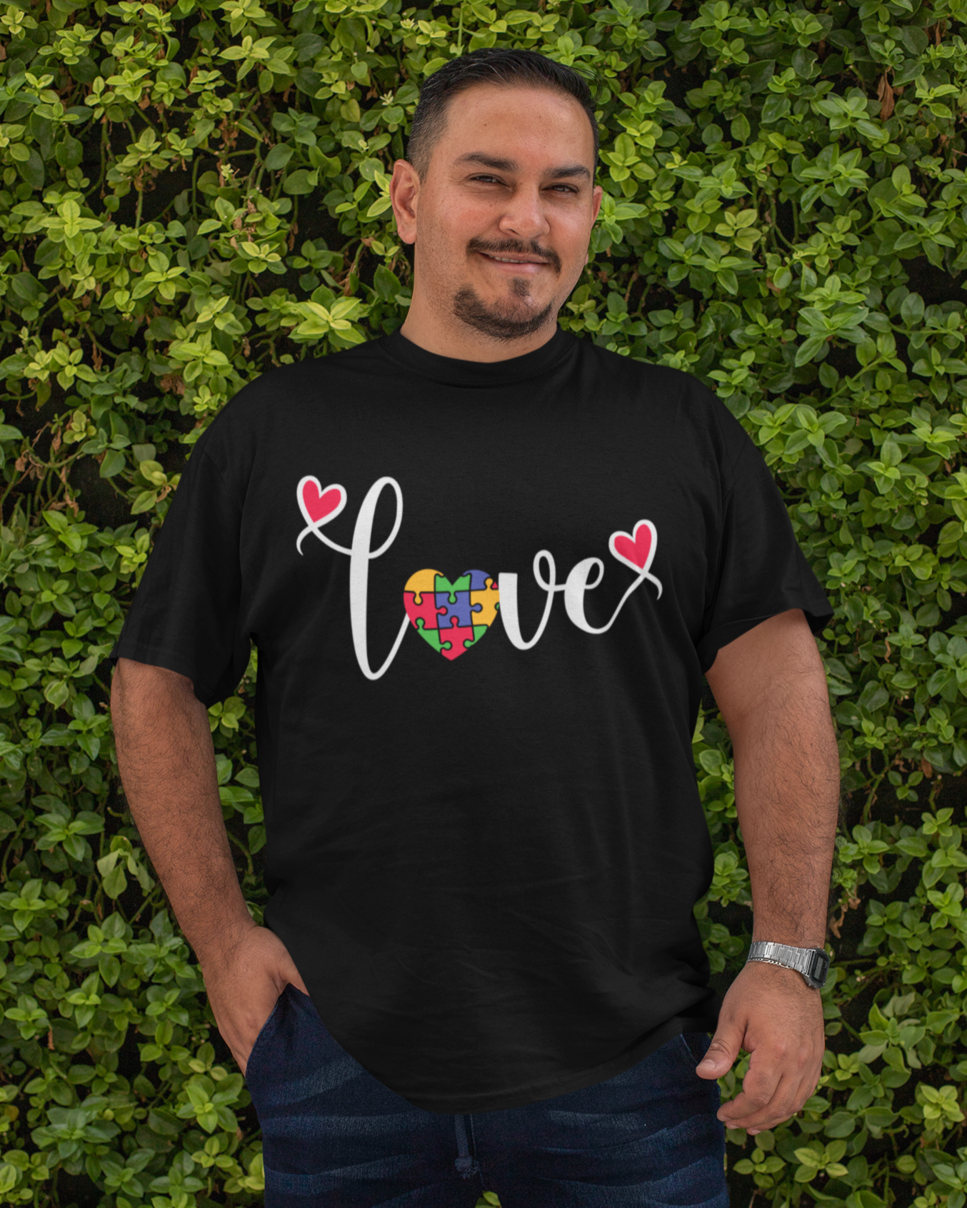 Άνδρας με μαύρο μπλουζάκι που γράφει "Love" με καρδιά-παζλ στο κέντρο, ποζάρει μπροστά σε πράσινο φόντο με φύλλα.