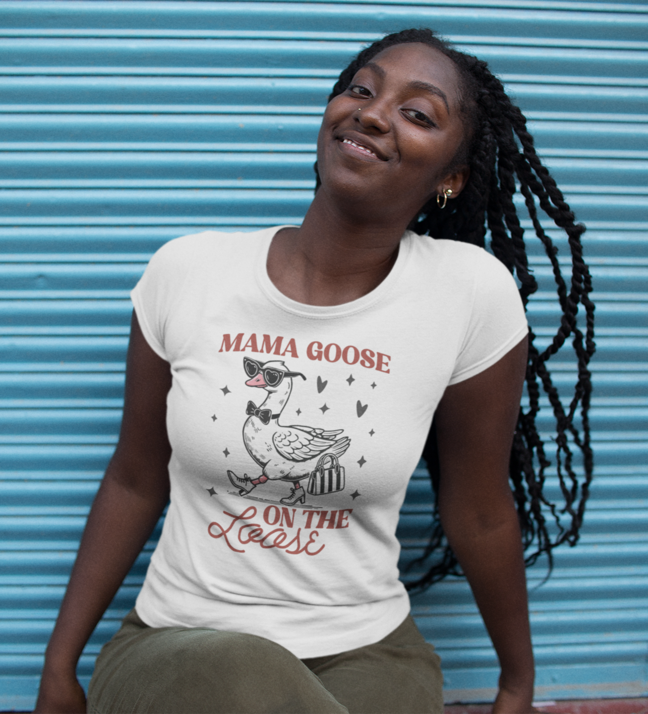 λευκό μπλουζάκι που γράφει "Mama Goose On The Loose" και σχέδιο χήνας με γυαλιά ηλίου και βαλίτσα, κάθεται μπροστά σε μπλε μεταλλικό φόντο.