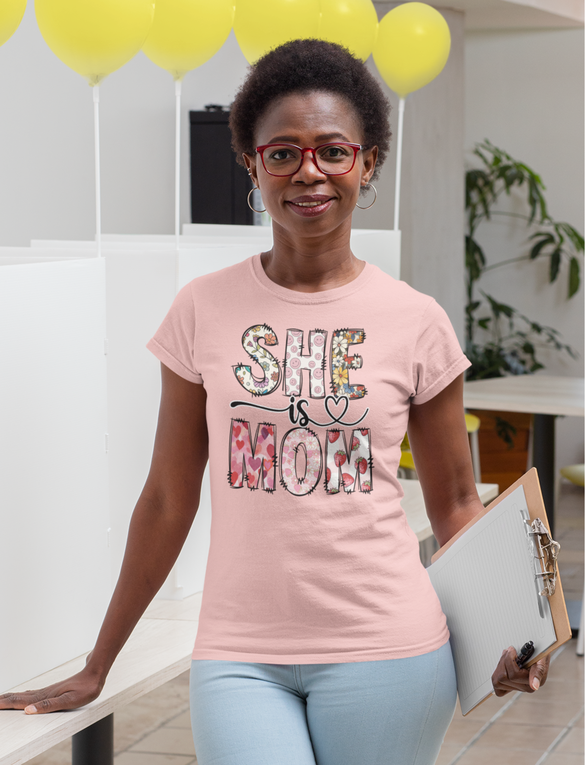ροζ μπλουζάκι που γράφει "She is Mom" με floral σχέδιο, κρατά clipboard και βρίσκεται σε χώρο με κίτρινα μπαλόνια.