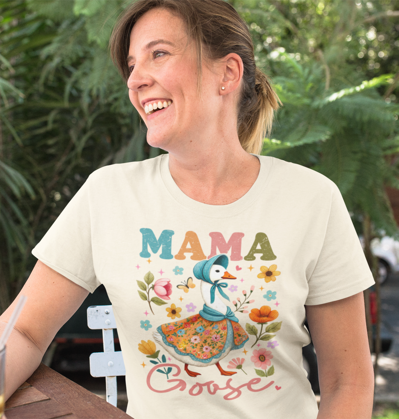 μπεζ μπλουζάκι που γράφει "MAMA Goose" με σχέδιο χήνα με φόρεμα και λουλούδια, κάθεται σε εξωτερικό καφέ και χαμογελά.