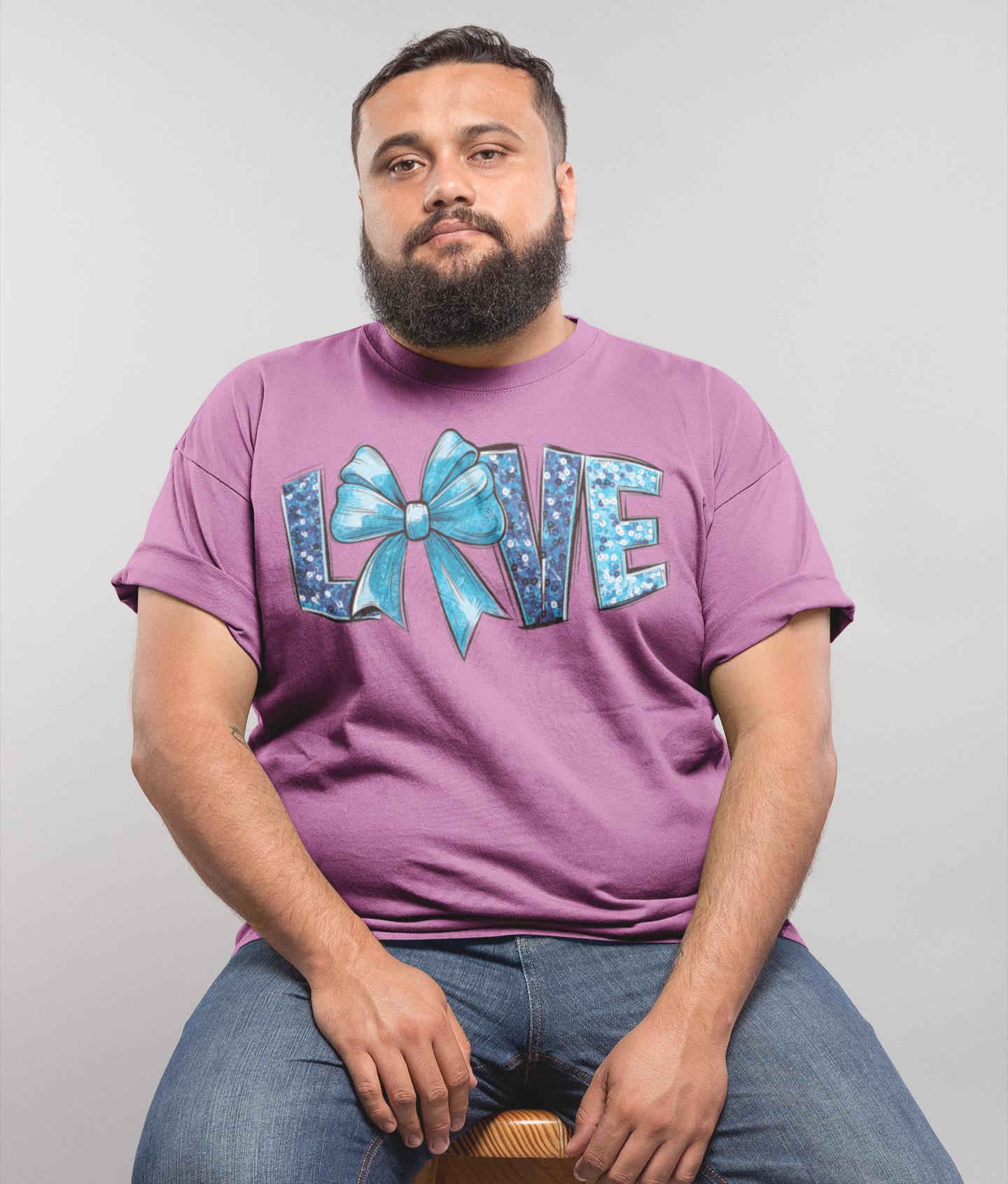 Άνδρας με μωβ μπλουζάκι plus size με σχέδιο LOVE και μπλε φιόγκο, κάθεται σε ξύλινο σκαμπό μπροστά από γκρι φόντο.
