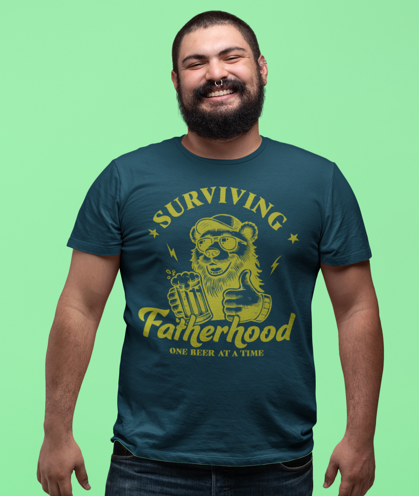 Γελαστός άνδρας με μούσι και piercing στη μύτη φοράει μπλουζάκι “Surviving Fatherhood – One Beer at a Time” με σχέδιο αρκούδας που κρατάει μπύρα.