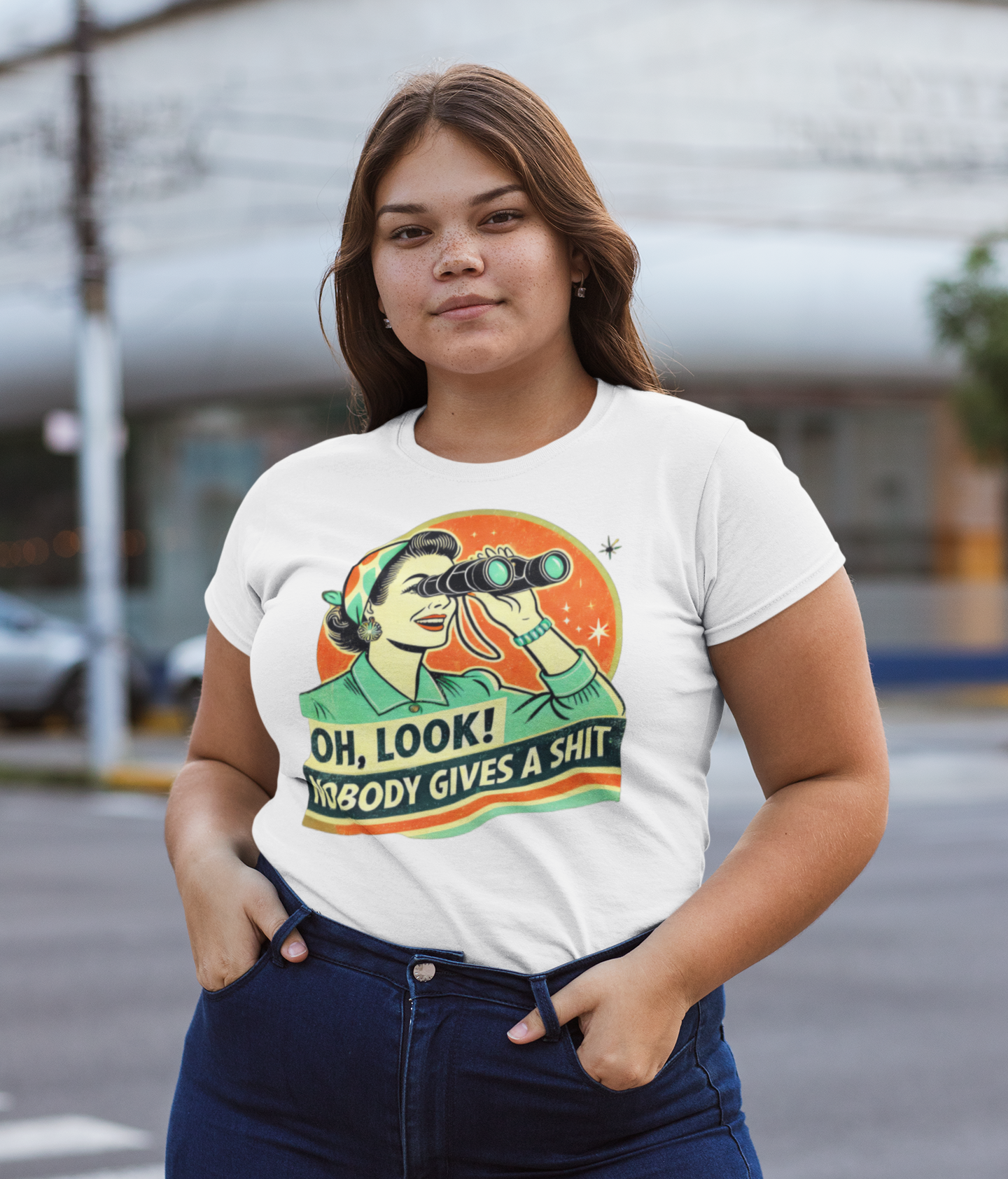 Γυναίκα φοράει λευκό t-shirt με ρετρό γραφικό μιας γυναίκας με κιάλια και το μήνυμα “Oh Look! Nobody Gives a Shit”, σε αστικό φόντο