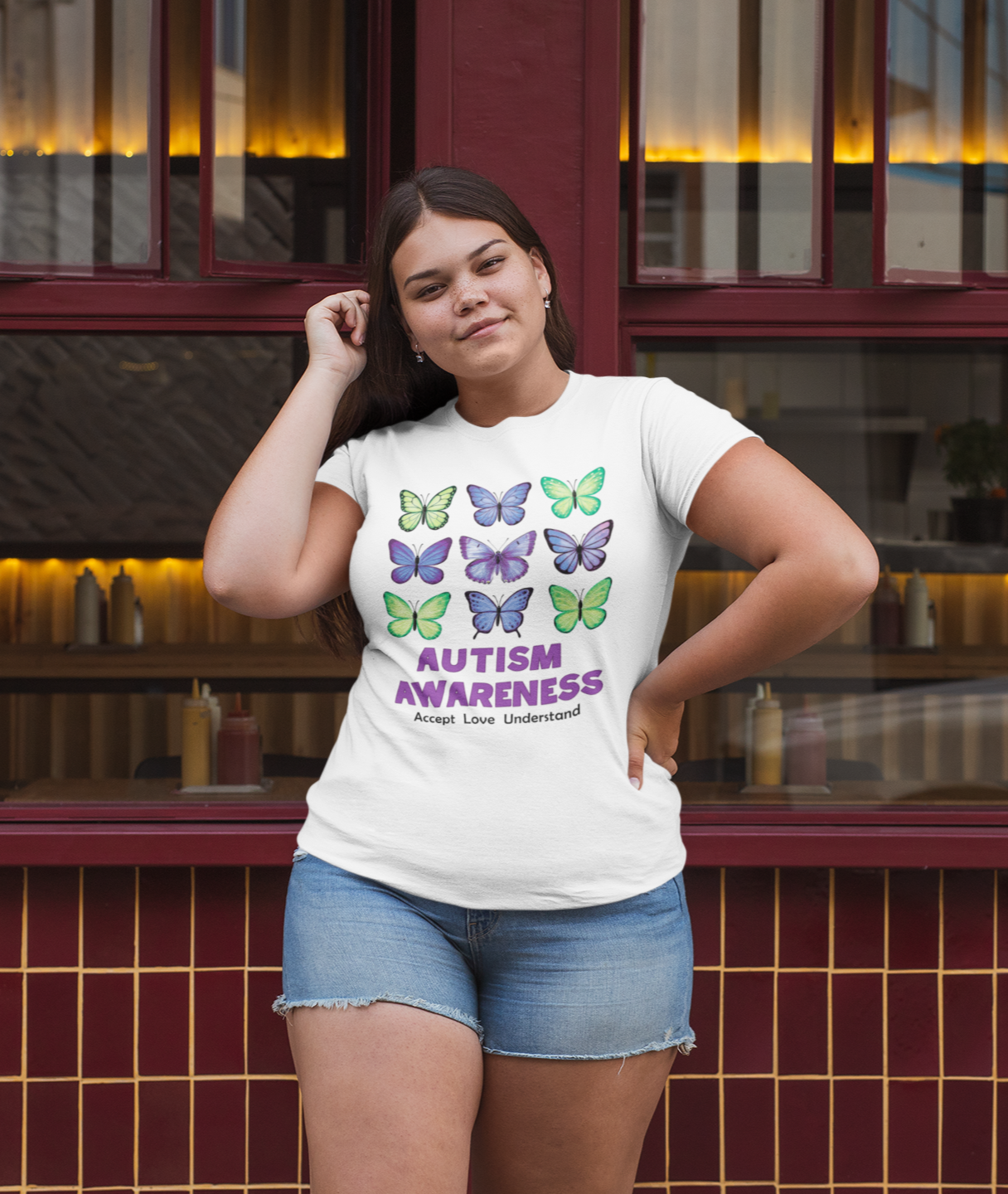 Γυναίκα με λευκό μπλουζάκι plus size με σχέδιο πολύχρωμες πεταλούδες και μήνυμα "Autism Awareness", ποζάρει έξω από εστιατόριο με κόκκινα πλαίσια.