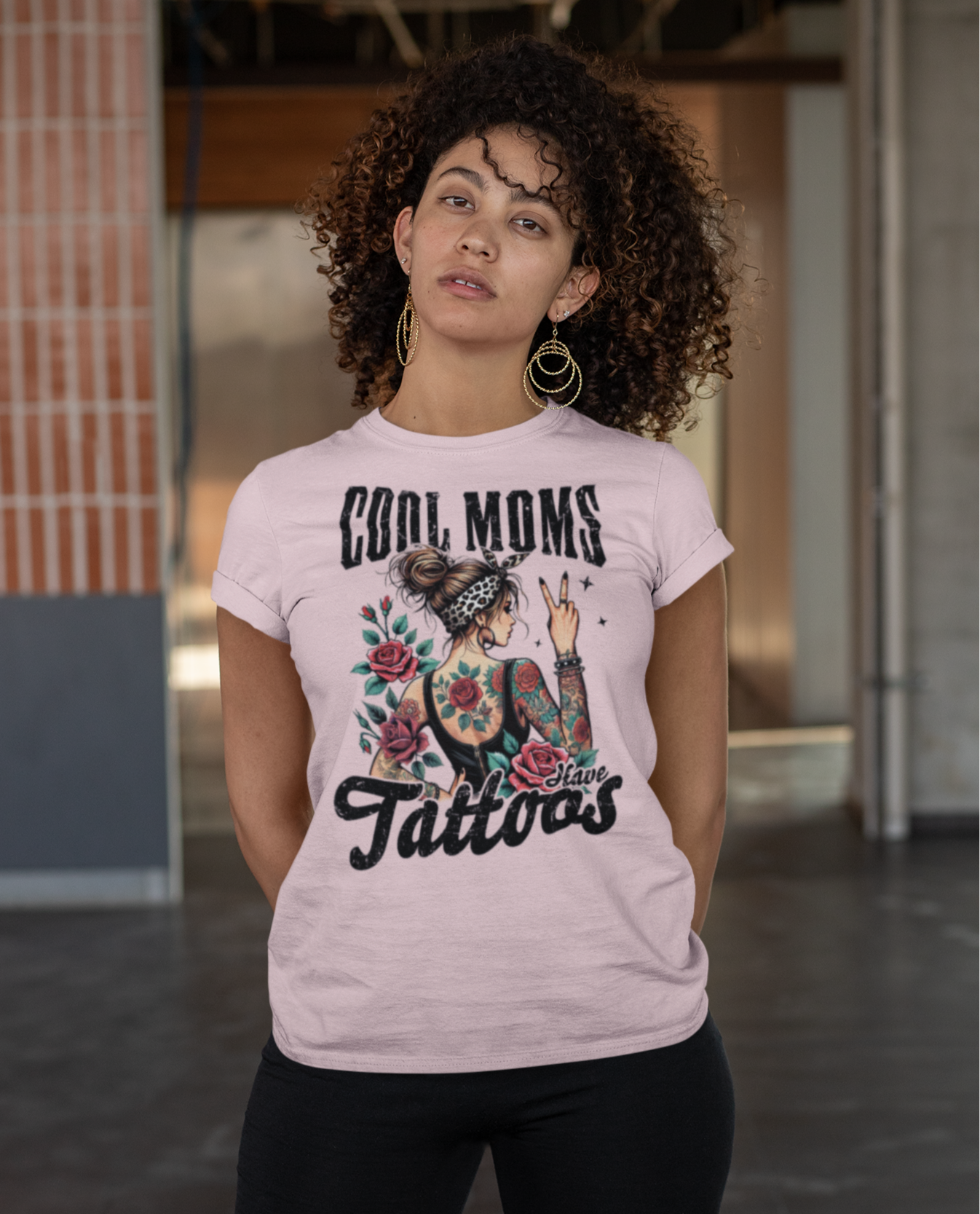 Γυναίκα με σγουρά μαλλιά φοράει ροζ μπλουζάκι με σχέδιο γυναίκας με τατουάζ και φράση “Cool Moms Have Tattoos”