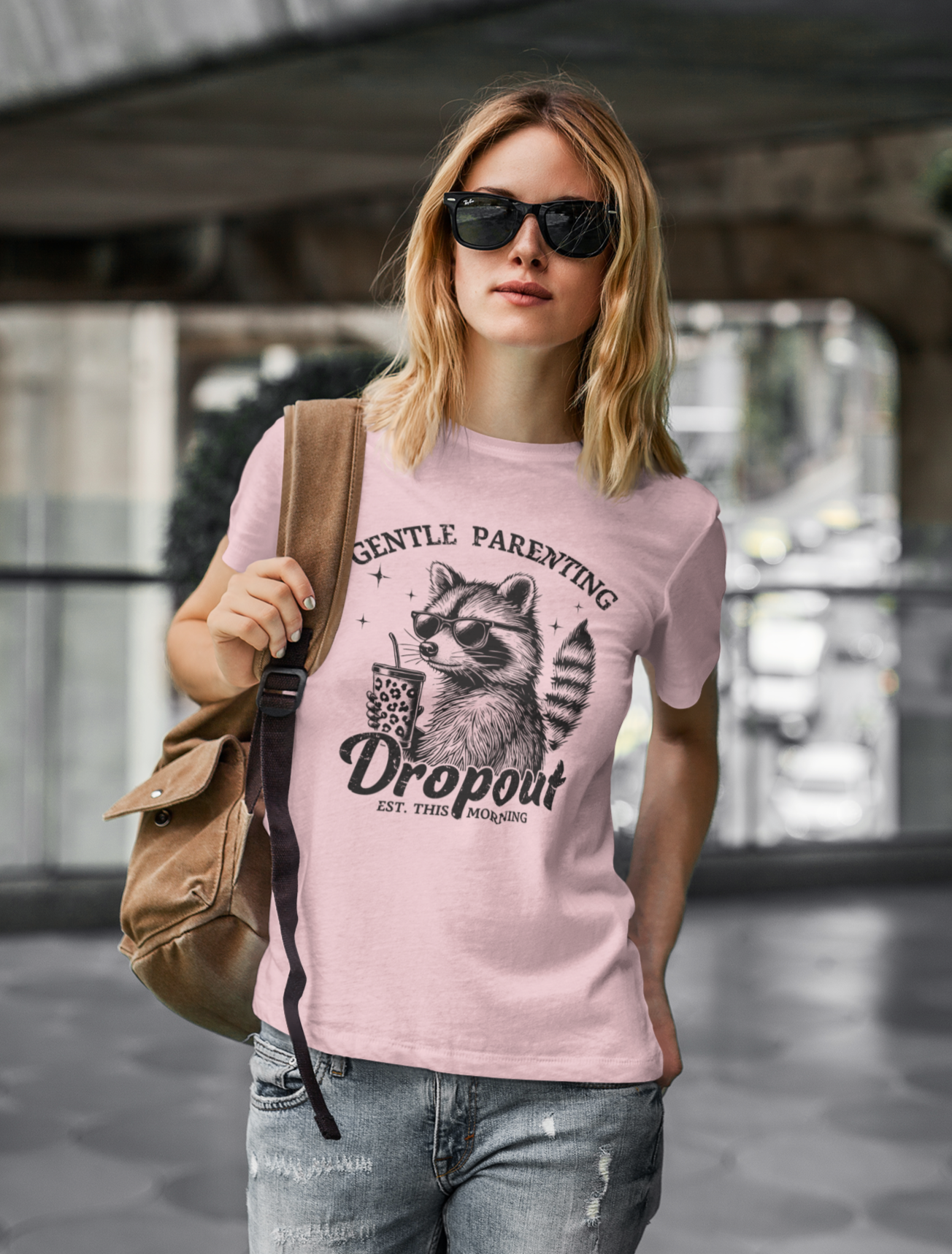 Γυναίκα με ροζ μπλουζάκι "Gentle Parenting Dropout" και σχέδιο ρακούν, γυαλιά ηλίου και σακίδιο, περπατά σε αστικό περιβάλλον.