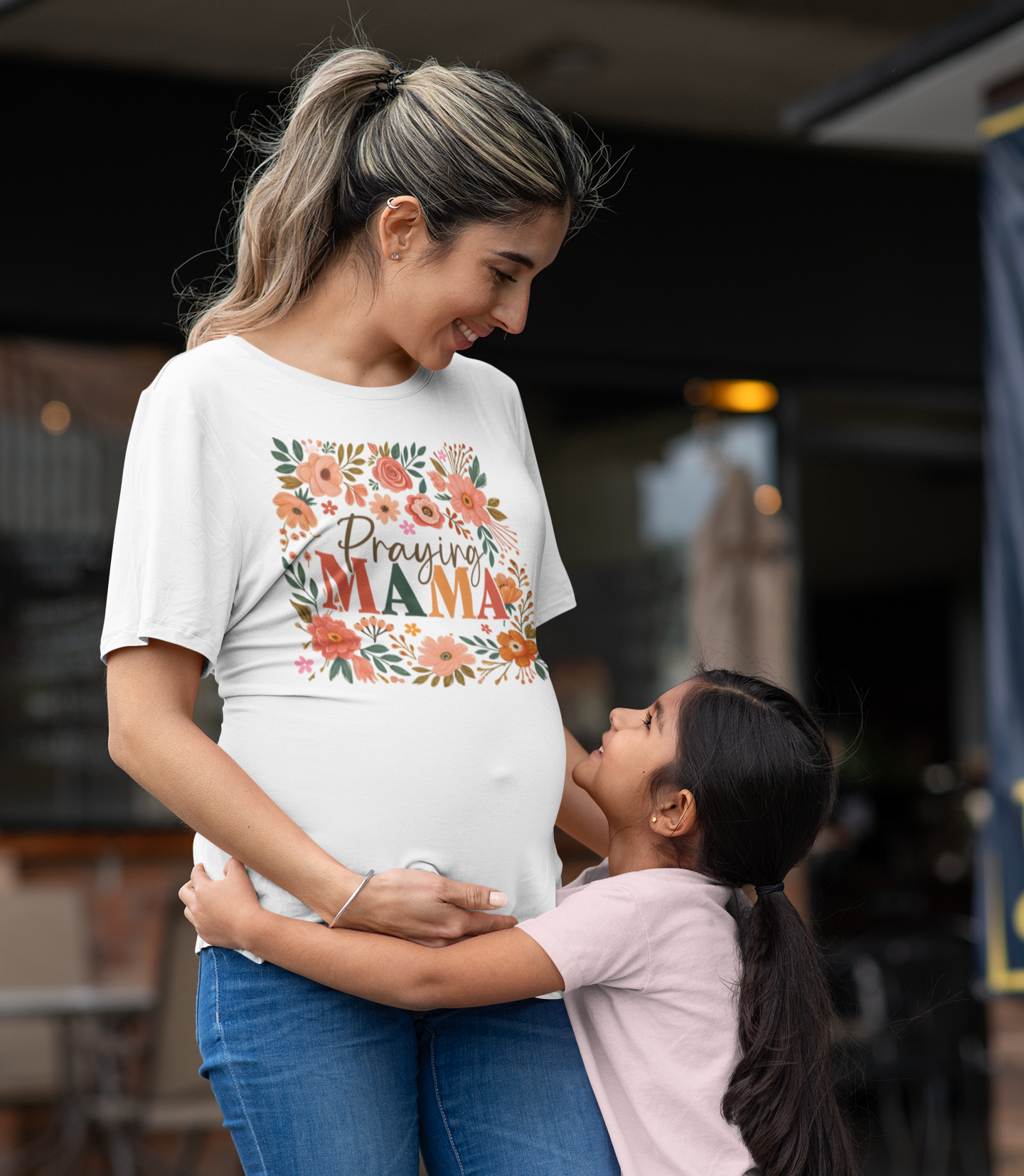 Έγκυος γυναίκα με λευκό μπλουζάκι που γράφει "Praying Mama" με λουλούδια, αγκαλιάζει και χαμογελά στην κόρη της σε εξωτερικό χώρο.