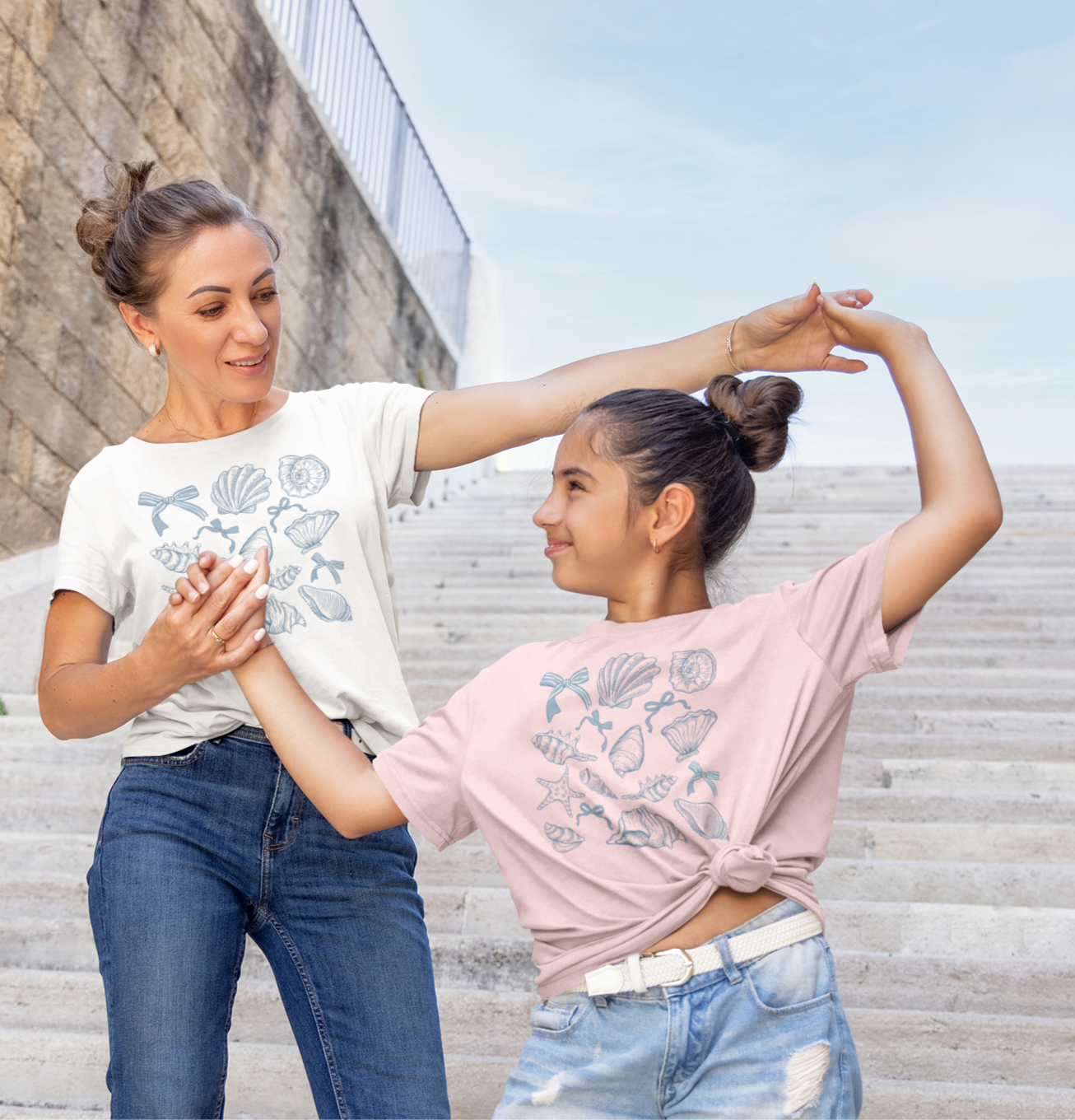 Μητέρα και κόρη φορούν μπλουζάκια με σχέδιο κοχύλια και ποζάρουν χαμογελαστές σε σκαλιά εξωτερικού χώρου, η μαμά με λευκό και η κόρη με ροζ μπλουζάκι.