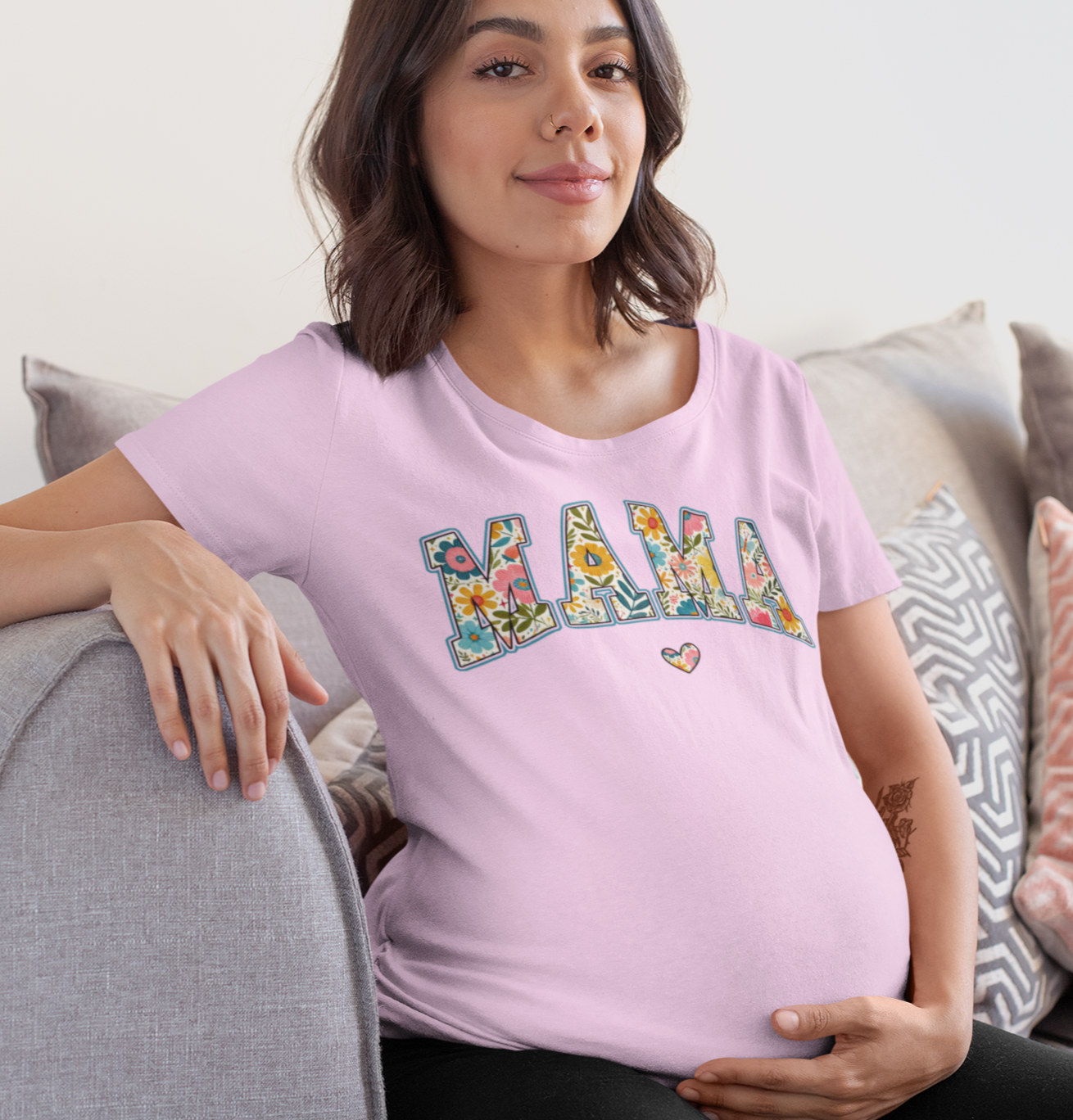 ροζ μπλουζάκι που γράφει MAMA με λουλουδάτο σχέδιο, κάθεται χαλαρή σε γκρι καναπέ με μαξιλάρια.