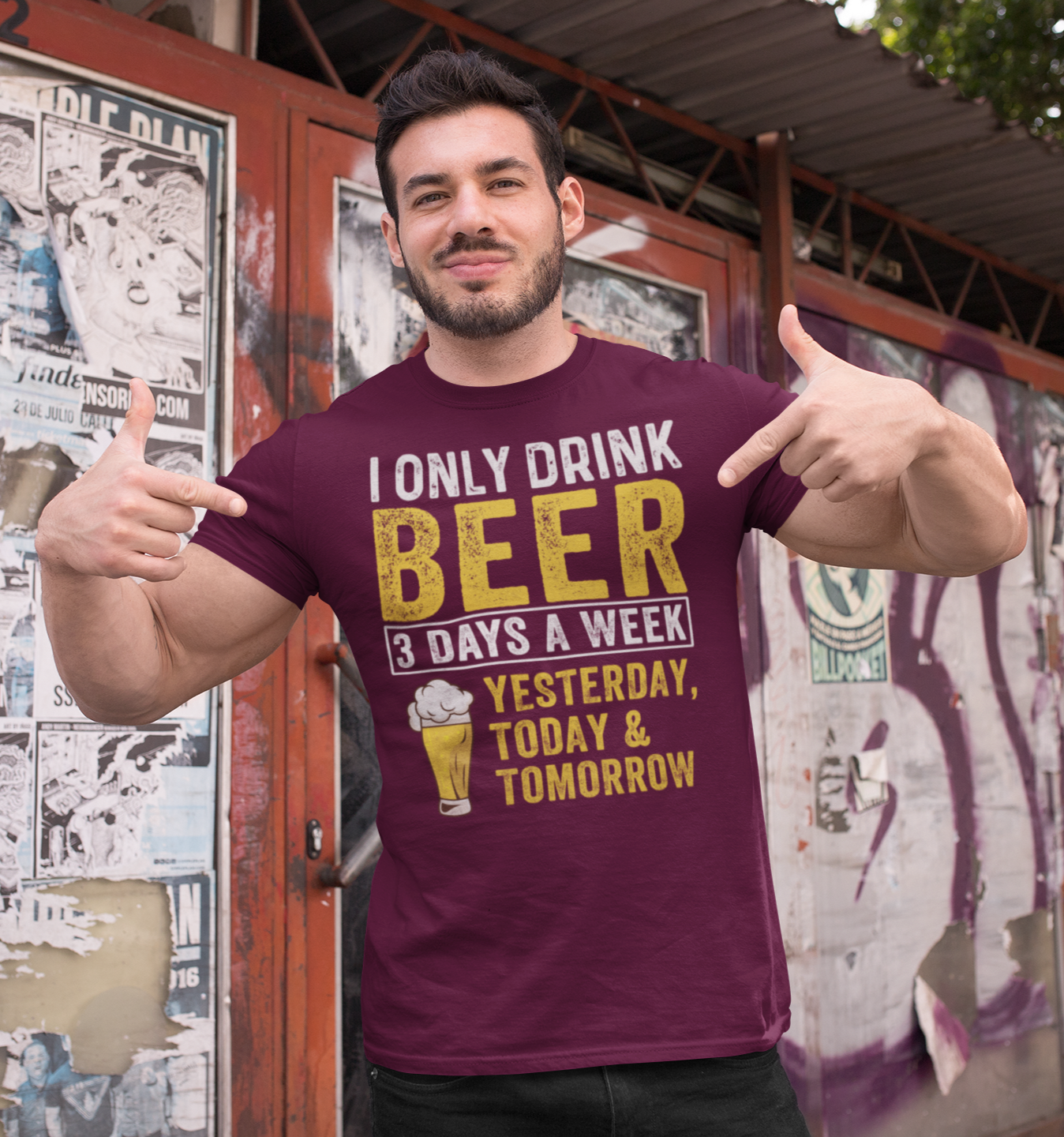 Γυμνασμένος άντρας δείχνει περήφανα το μπορντό μπλουζάκι του με μήνυμα “I Only Drink Beer 3 Days a Week – Yesterday, Today & Tomorrow” μπροστά σε τοίχο με αφίσες.