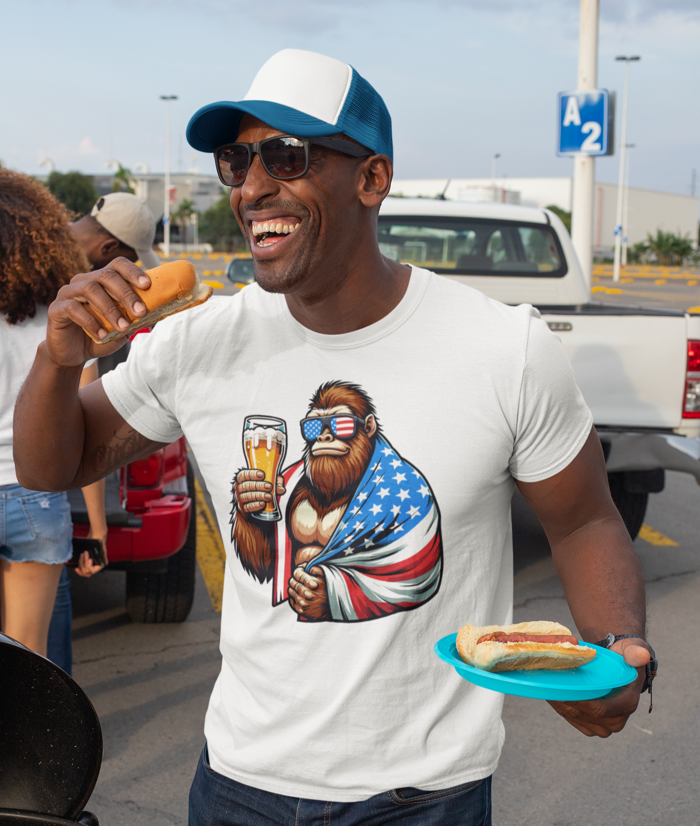 Άνδρας με καπέλο φορτηγού και γυαλιά ηλίου γελάει και κρατά hot dog φορώντας t-shirt με σχέδιο Bigfoot που κρατά μπύρα και είναι τυλιγμένος με αμερικανική σημαία.