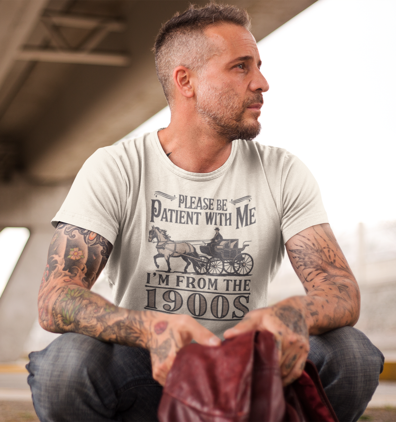 Άνδρας με τατουάζ κάθεται κάτω από γέφυρα φορώντας μπλουζάκι με το χιουμοριστικό μήνυμα "Please be patient with me – I'm from the 1900s" και σχέδιο άμαξας.