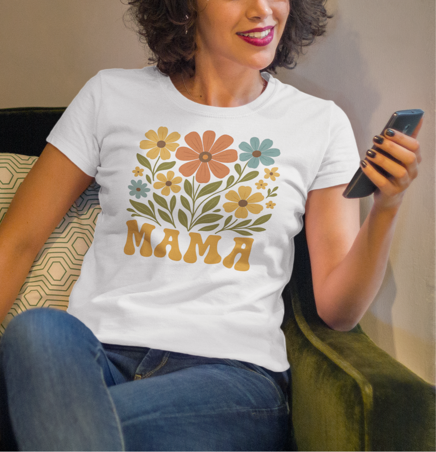 Γυναίκα με λευκό μπλουζάκι που γράφει "MAMA" με λουλούδια, κάθεται σε πράσινη πολυθρόνα και κρατά κινητό, χαμογελώντας.