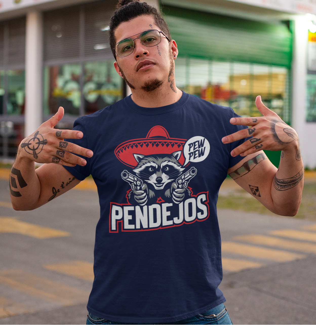 Άντρας με τατουάζ φοράει μπλε μπλουζάκι με ρακούν που κρατάει όπλα και τη φράση “Pew Pew Pendejos”.