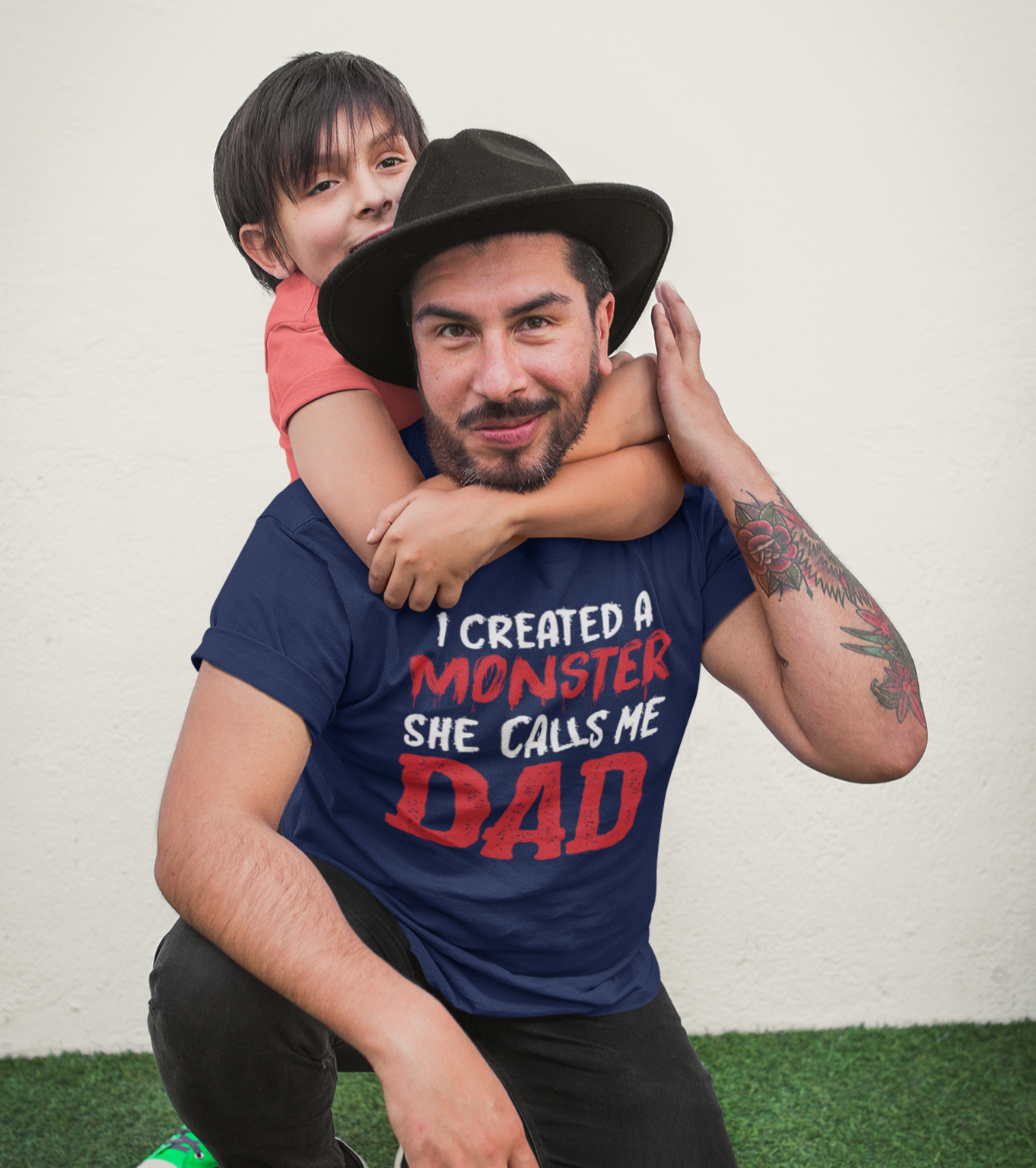 Μπαμπάς με τατουάζ και καπέλο γονατιστός, αγκαλιάζει την κόρη του που τον αγκαλιάζει από πίσω. Φοράει μπλουζάκι με χιουμοριστικό μήνυμα: “I Created a Monster, She Calls Me Dad”.