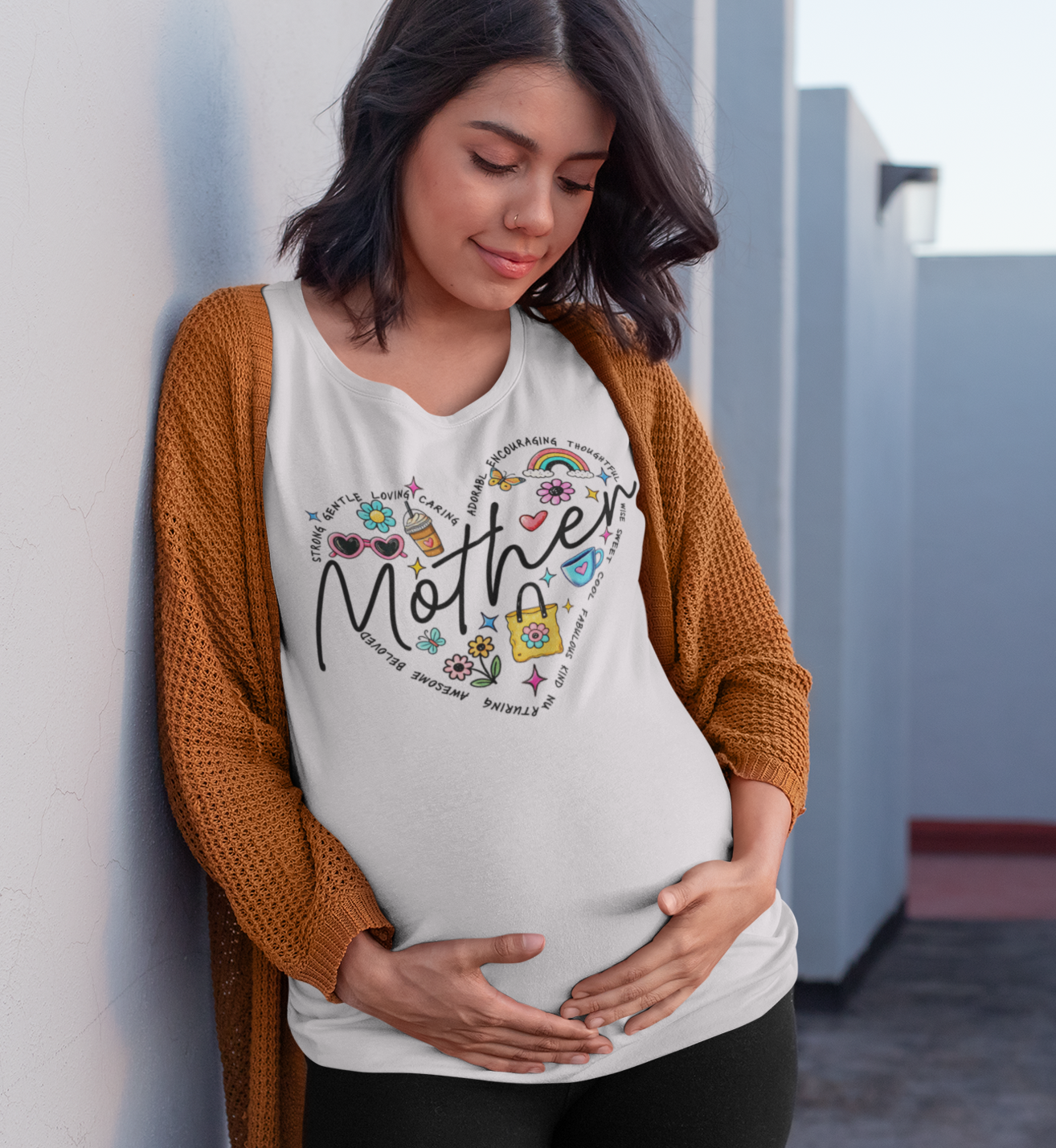  λευκό μπλουζάκι που γράφει "Mother" με σχέδιο καρδιά και λουλούδια, ποζάρει αγκαλιάζοντας την κοιλιά της, σε εξωτερικό χώρο με λευκούς τοίχους.