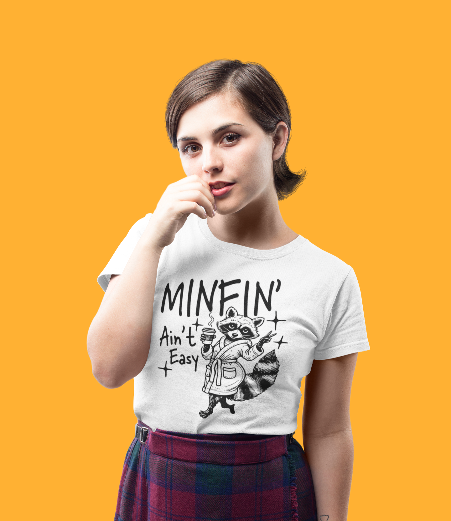 Νεαρή γυναίκα φοράει λευκό μπλουζάκι με ρακούν που κρατάει καφέ και γράφει “Minfin’ Ain’t Easy”, σε στούντιο με πορτοκαλί φόντο.
